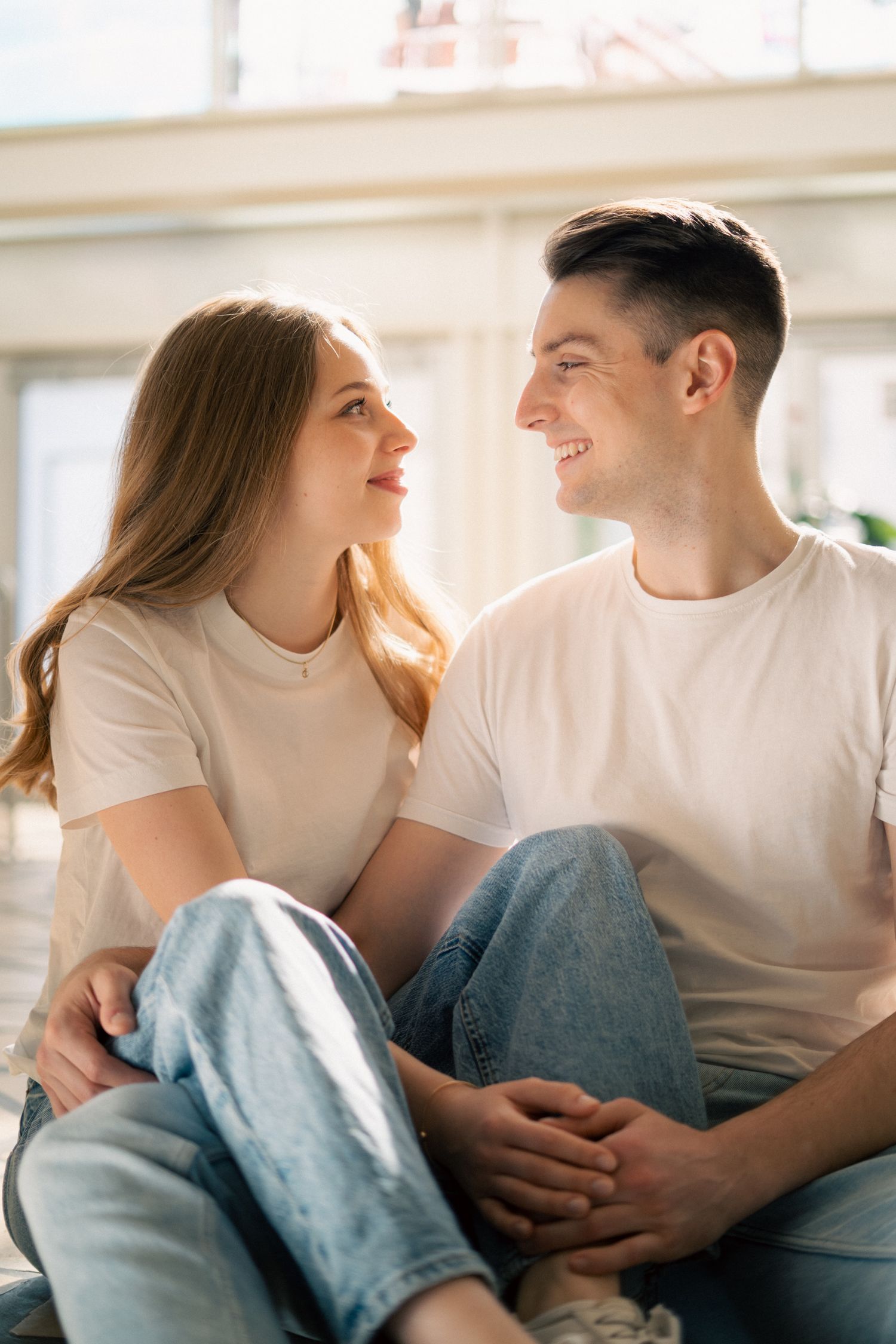 Happy young couple sitting on floor at home, smiling and looking at each other in casual white t-shirts and jeans.
