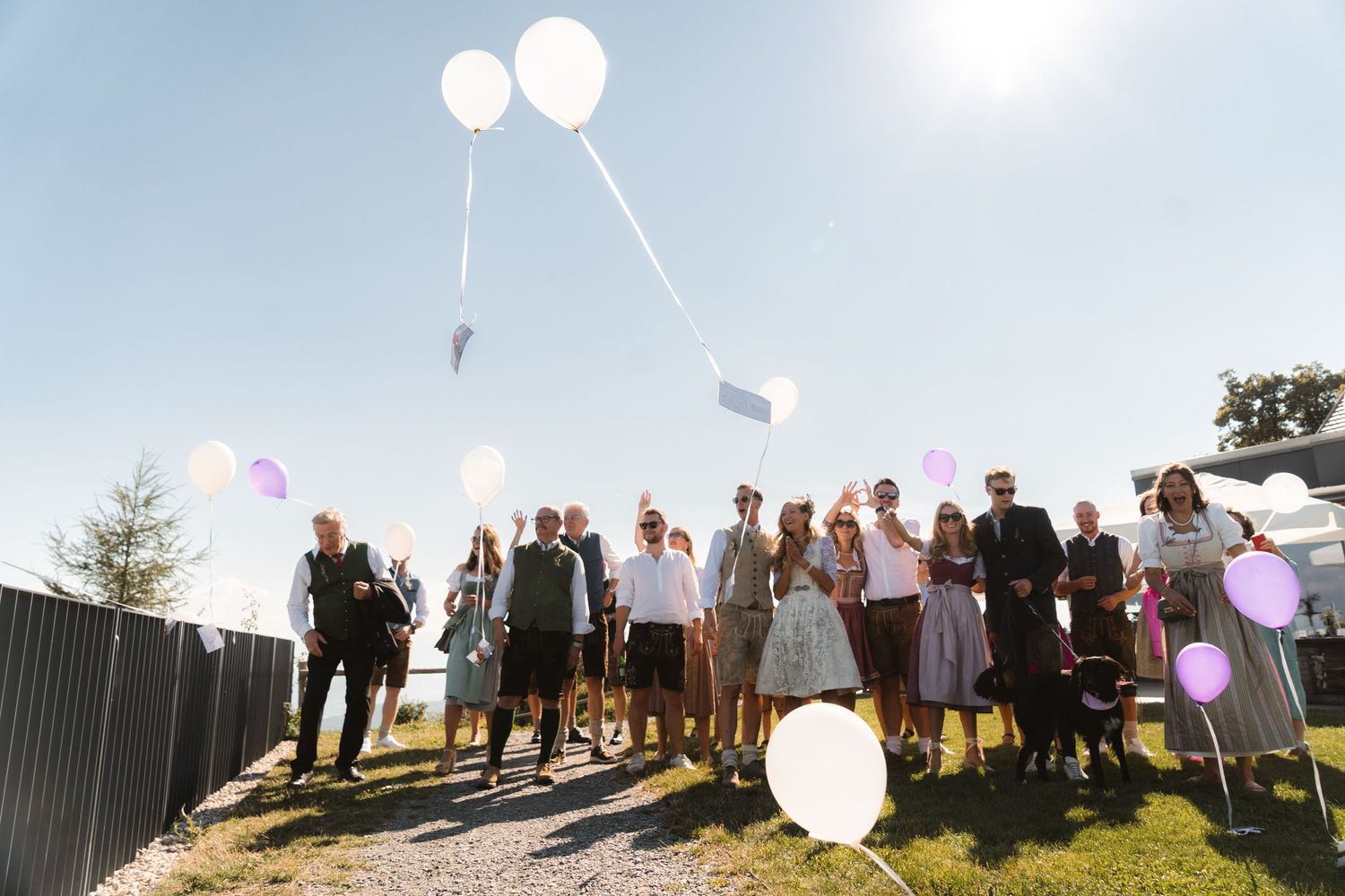 Wedding guests release white and purple balloons into a sunny blue sky during outdoor celebration.