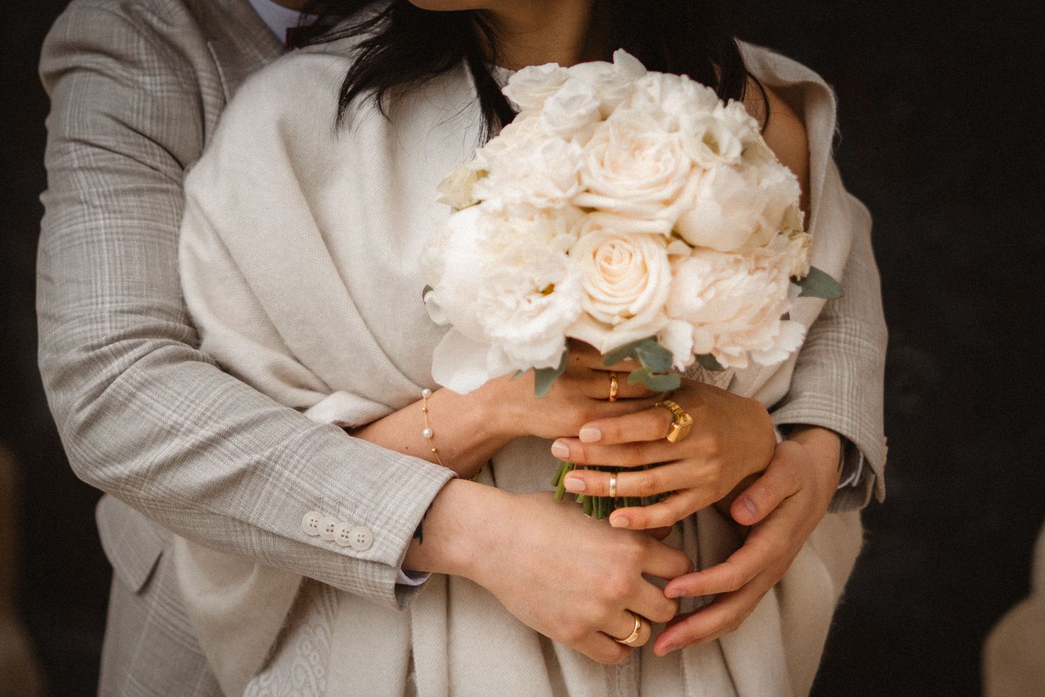 A couple embraces while holding a bouquet of white roses, with wedding rings visible on their hands.