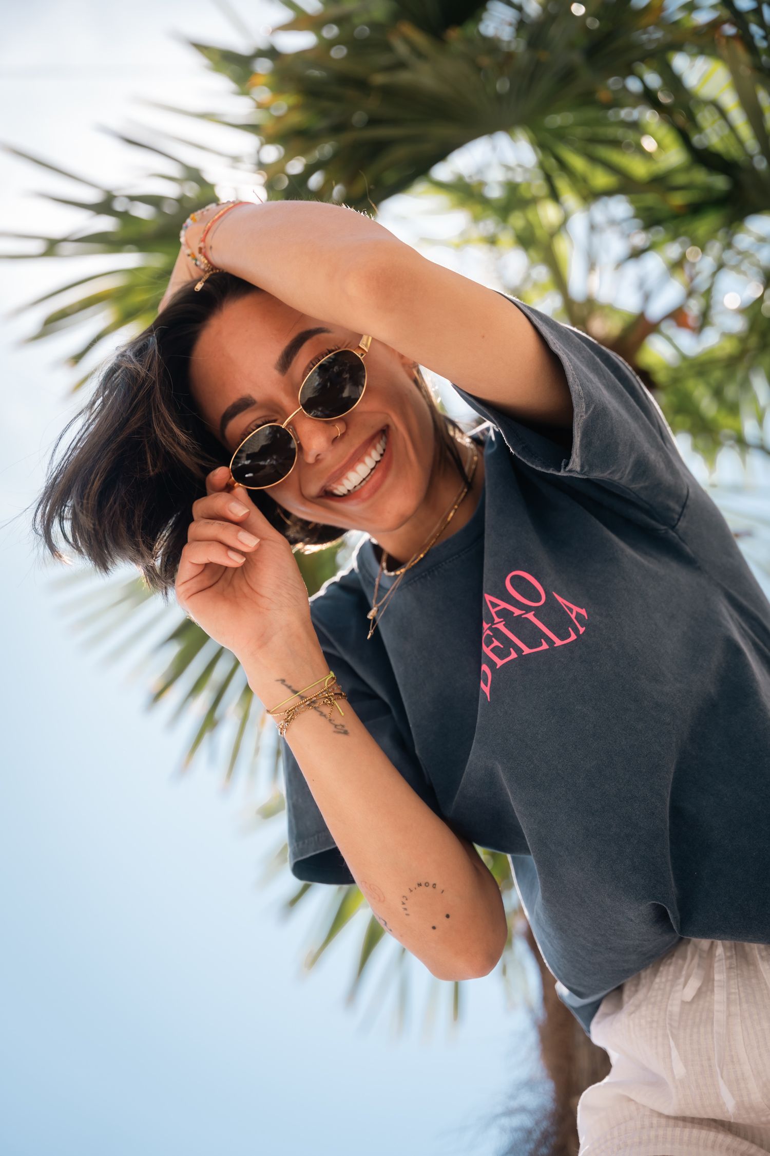Happy woman in gray graphic tee adjusting sunglasses while posing near tropical palm tree.
