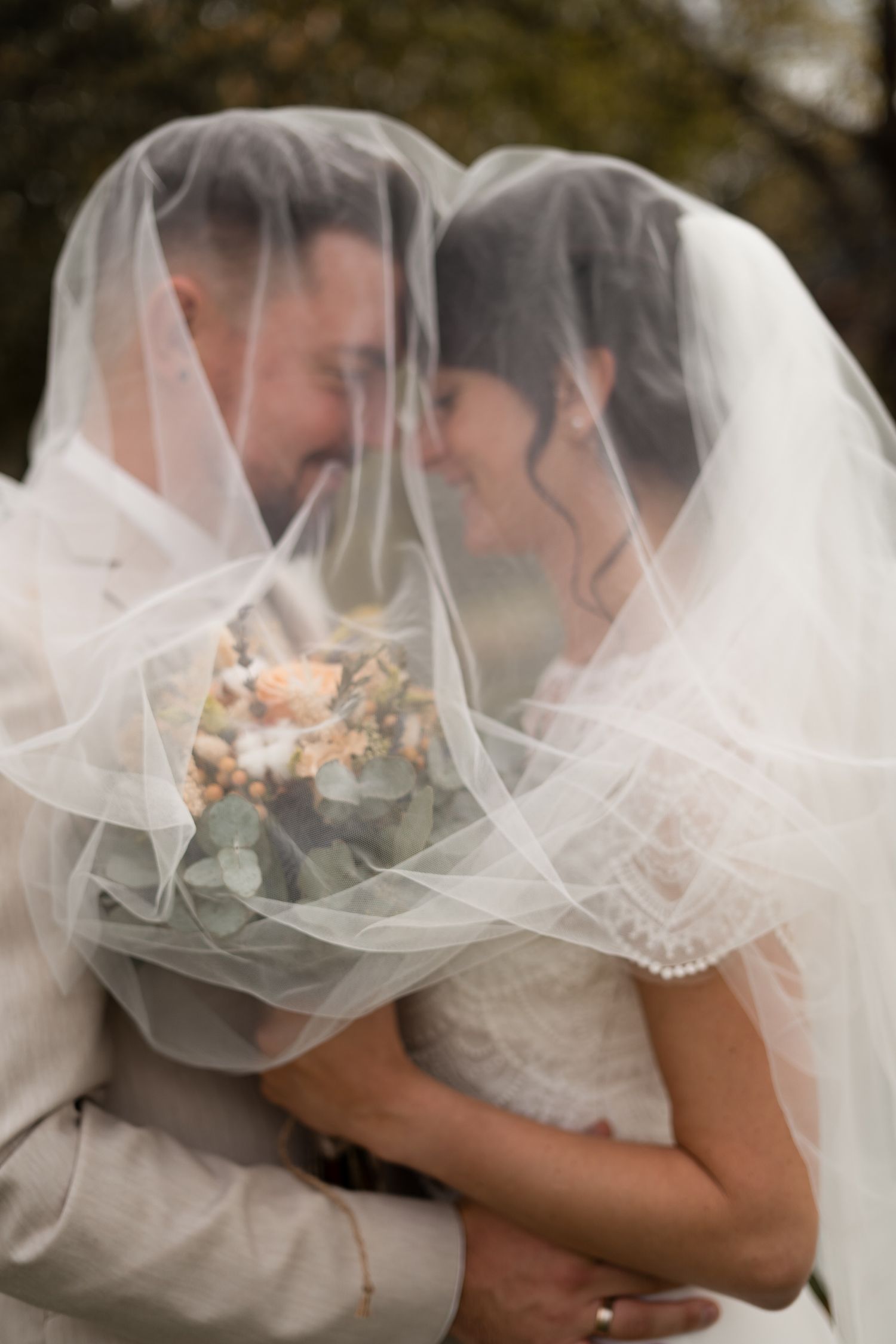 Romantic close-up of smiling bride and groom nearly kissing beneath flowing wedding veil holding floral bouquet.