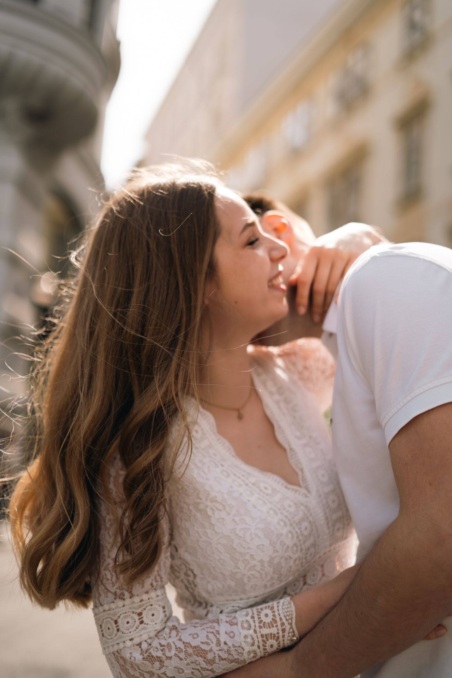 Romantic couple sharing a tender kiss outdoors in a sunlit European city, woman in white lace dress.
