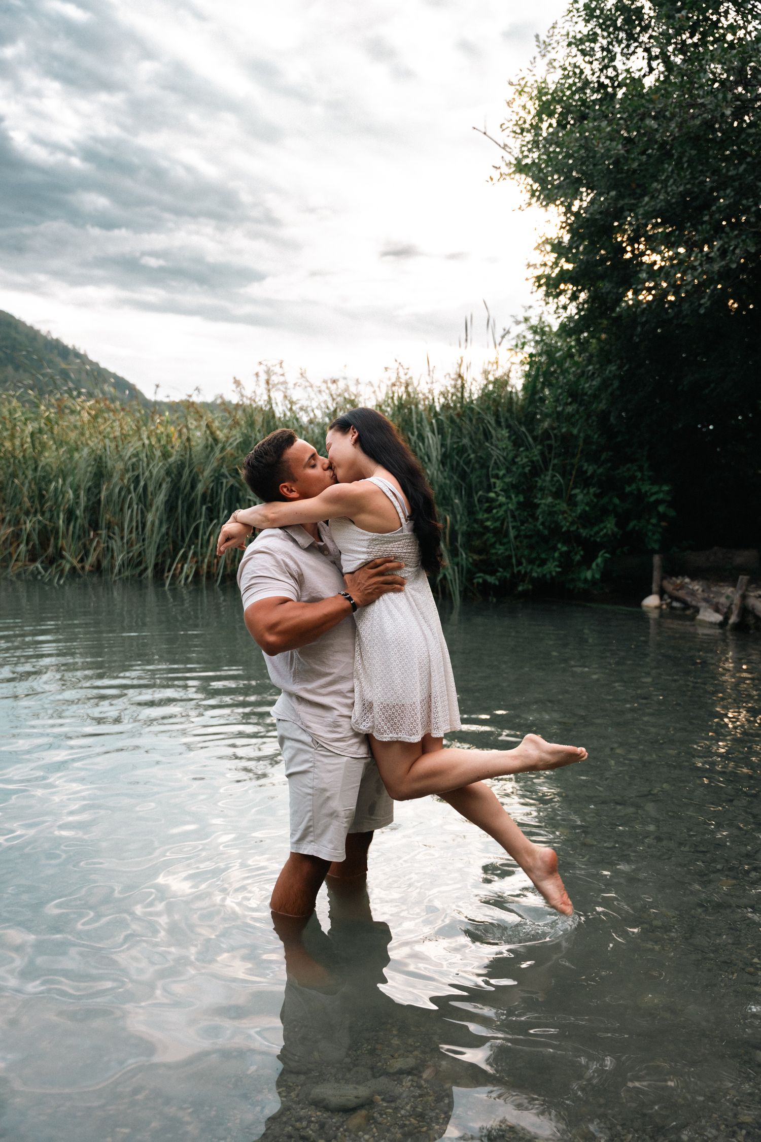 Couple sharing a kiss in shallow river water, man lifting woman in white dress outdoors.