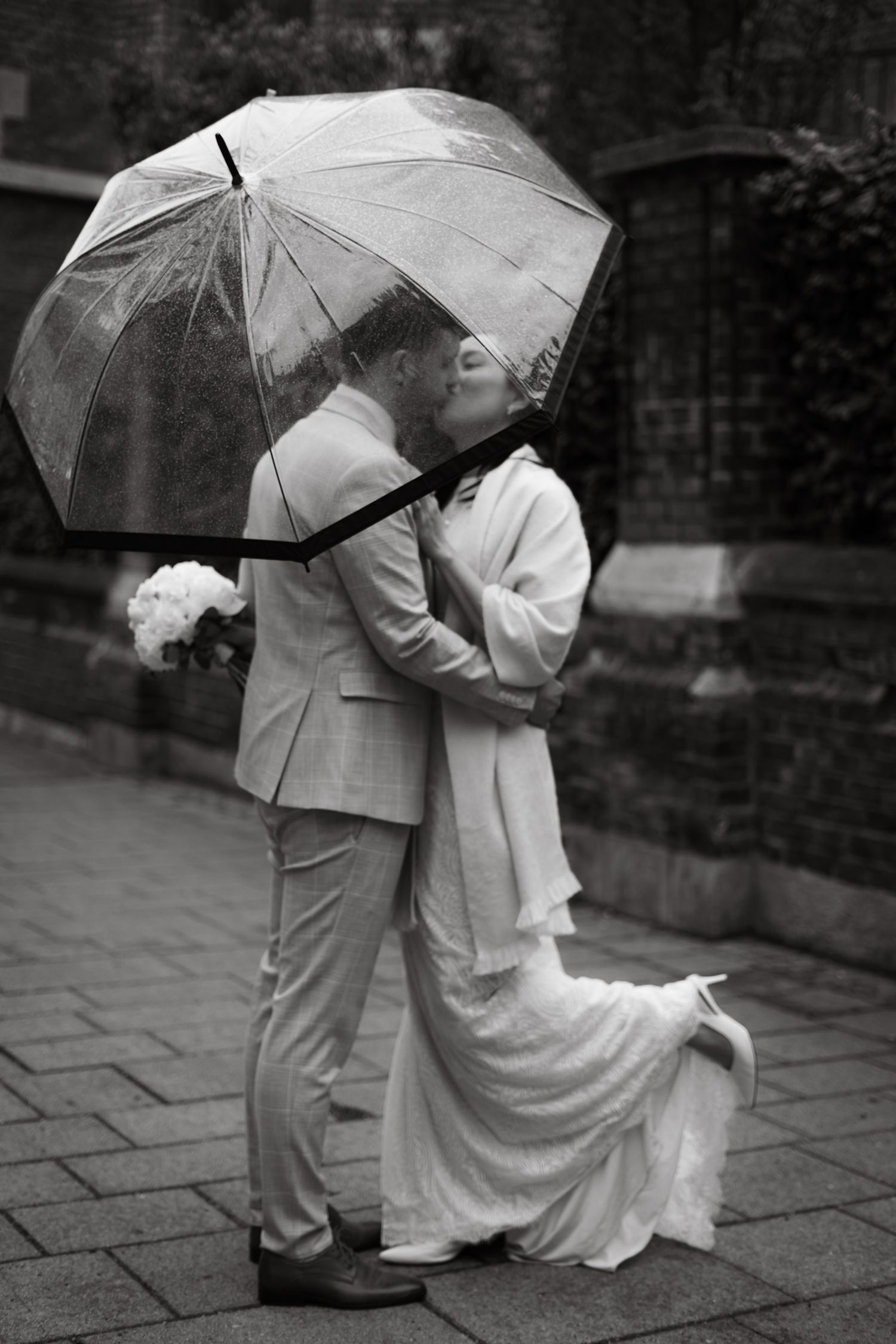 Newlyweds share a kiss under a clear umbrella in the rain in black and white.