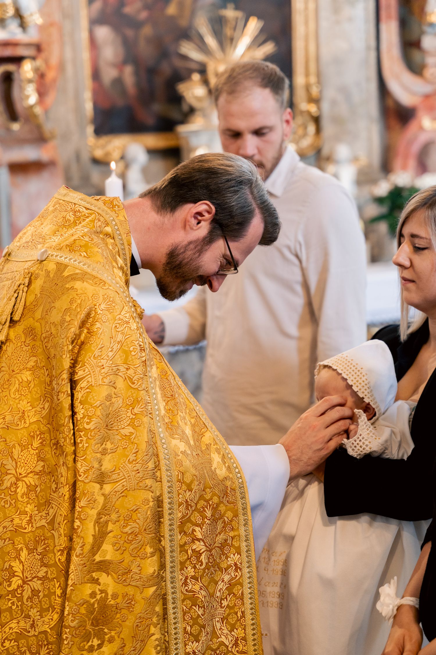 An Orthodox priest in golden robes performs a baptism ritual with a mother and infant.