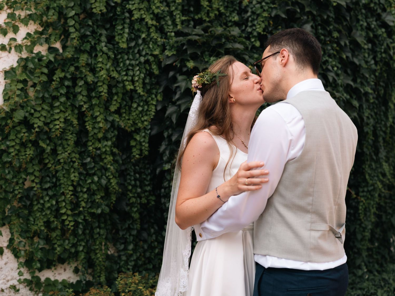 Bride and groom share a romantic kiss in front of a lush green ivy-covered wall on their wedding day.