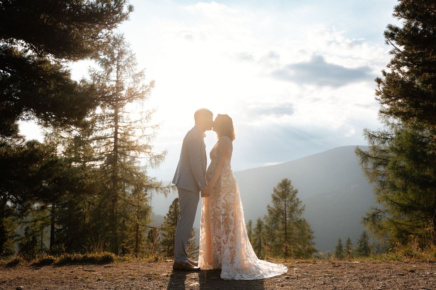 Couple shares romantic kiss at sunset in mountain forest wedding photo.