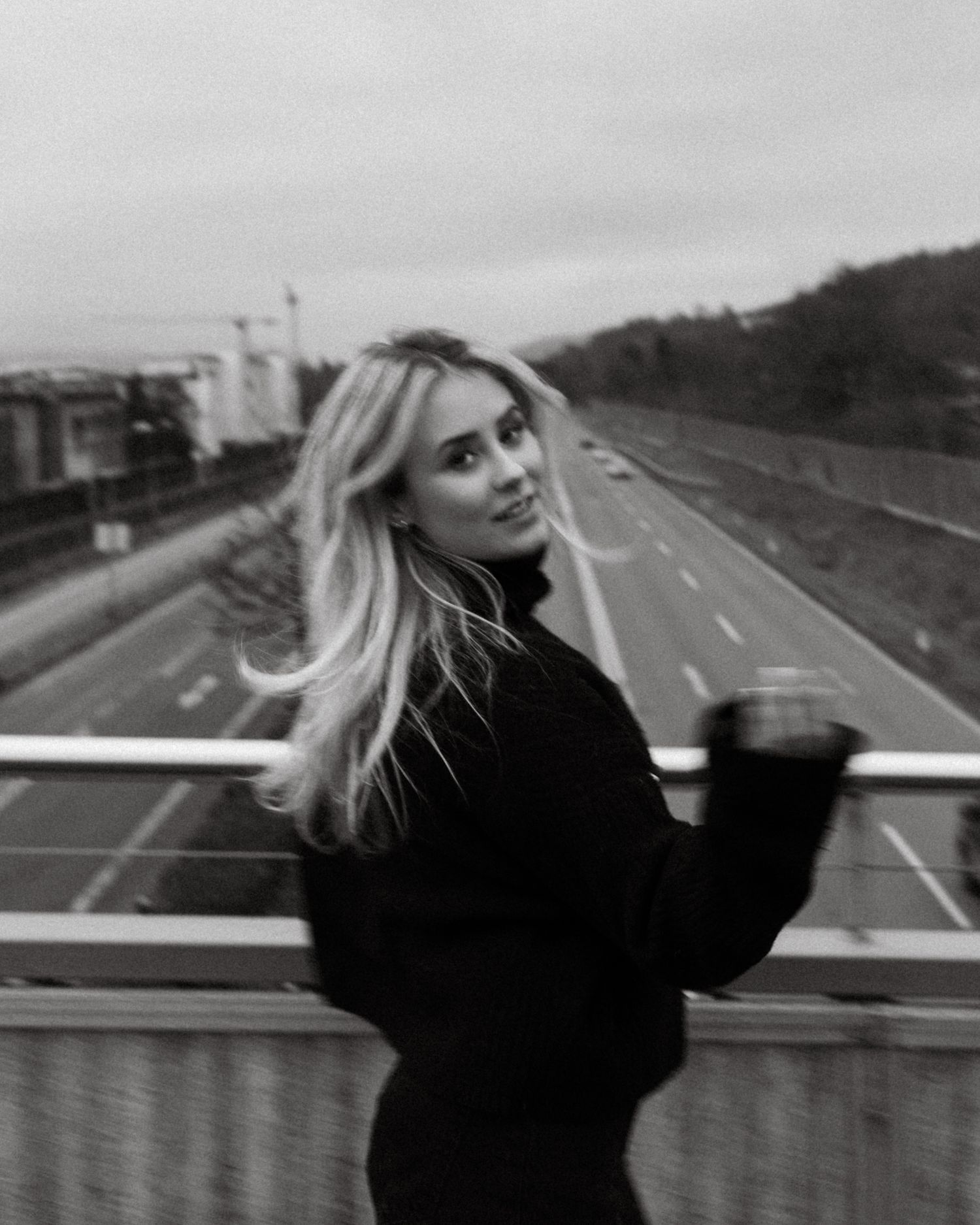 Black and white photo of blonde woman on highway overpass bridge smiling.