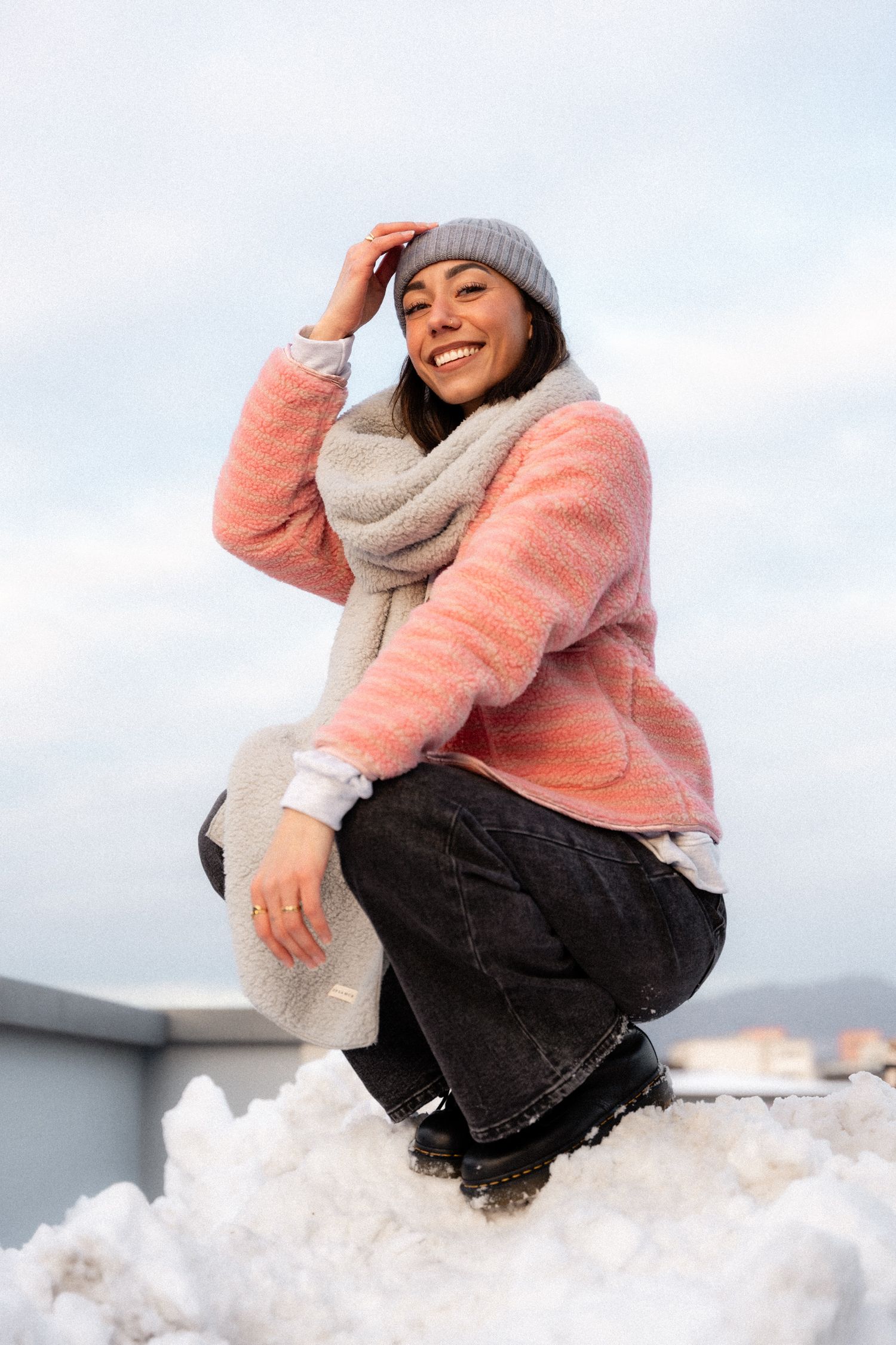 Smiling woman in a pink jacket, grey scarf, and beanie posing on a snow pile outdoors.