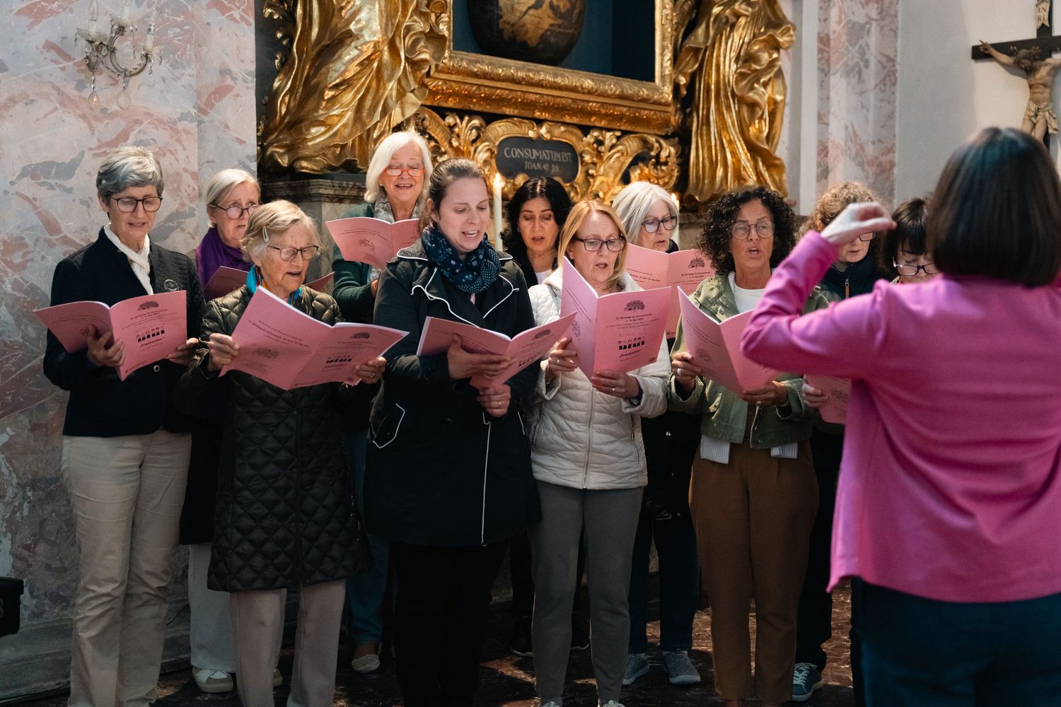 Women's choir performing with pink sheet music booklets led by a conductor in a decorated church.