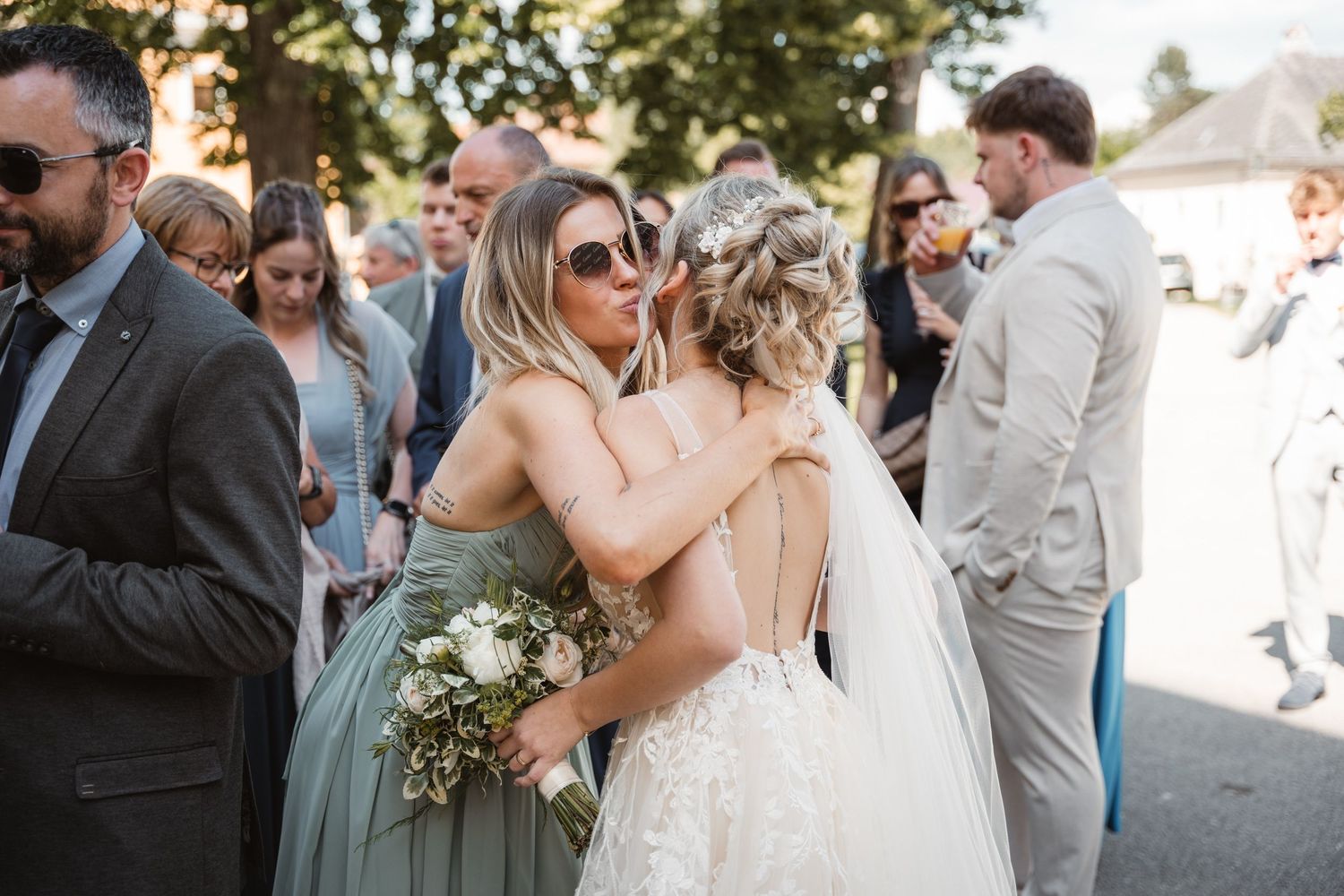 Bride in lace gown embracing a bridesmaid in sage green dress holding bouquet at outdoor wedding.