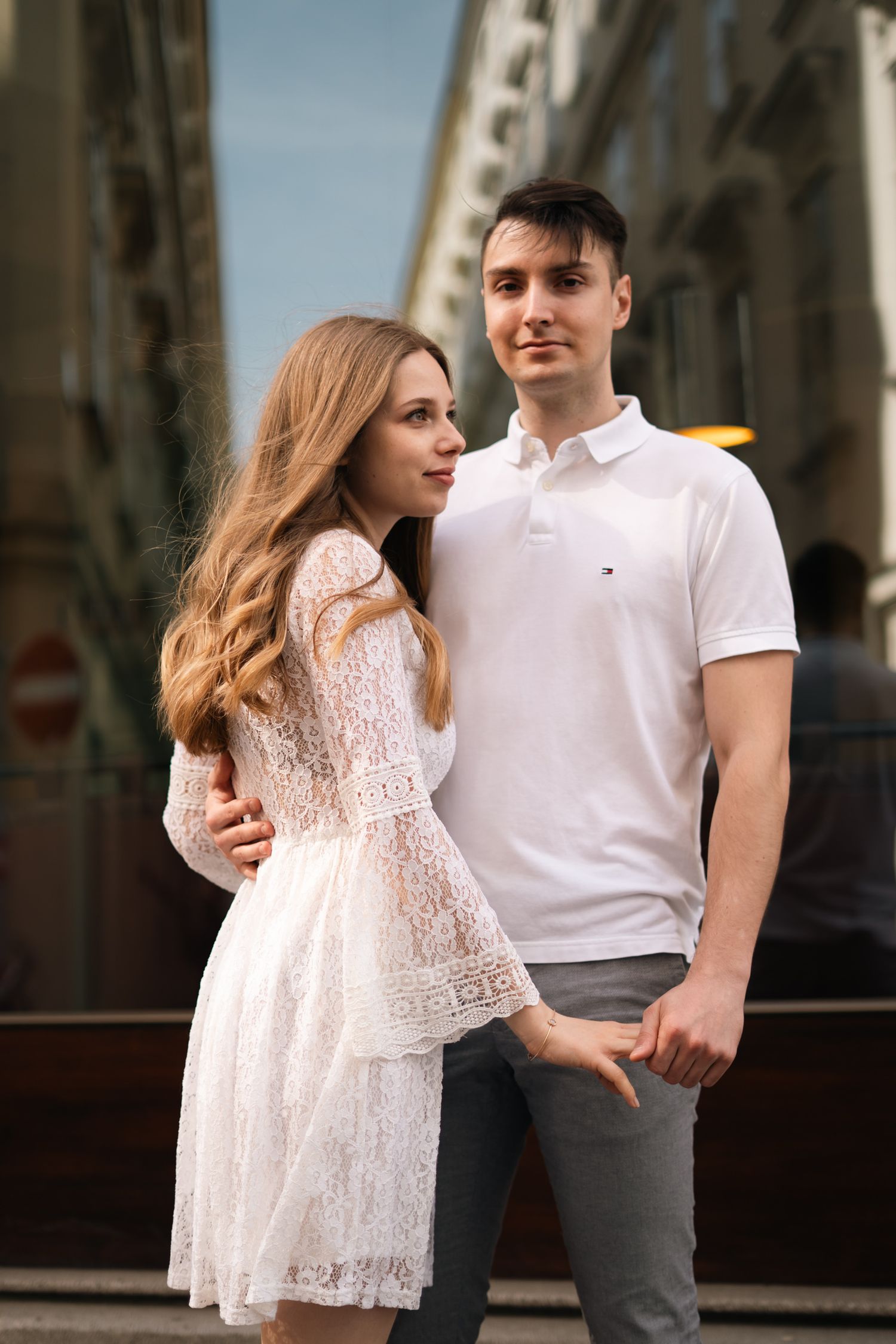 Young couple embracing on a sunny European city street, woman in white lace dress and man in white polo shirt.