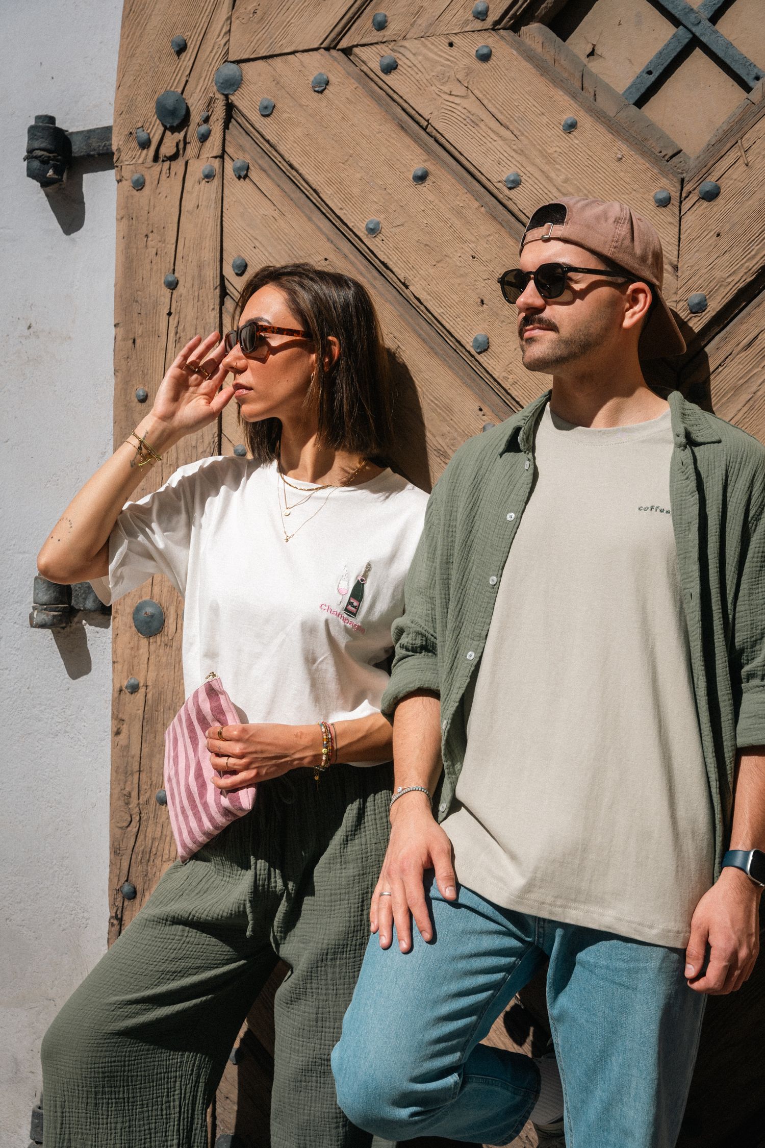 A stylish couple wearing casual streetwear stands against a rustic wooden door in sunny outdoor setting.