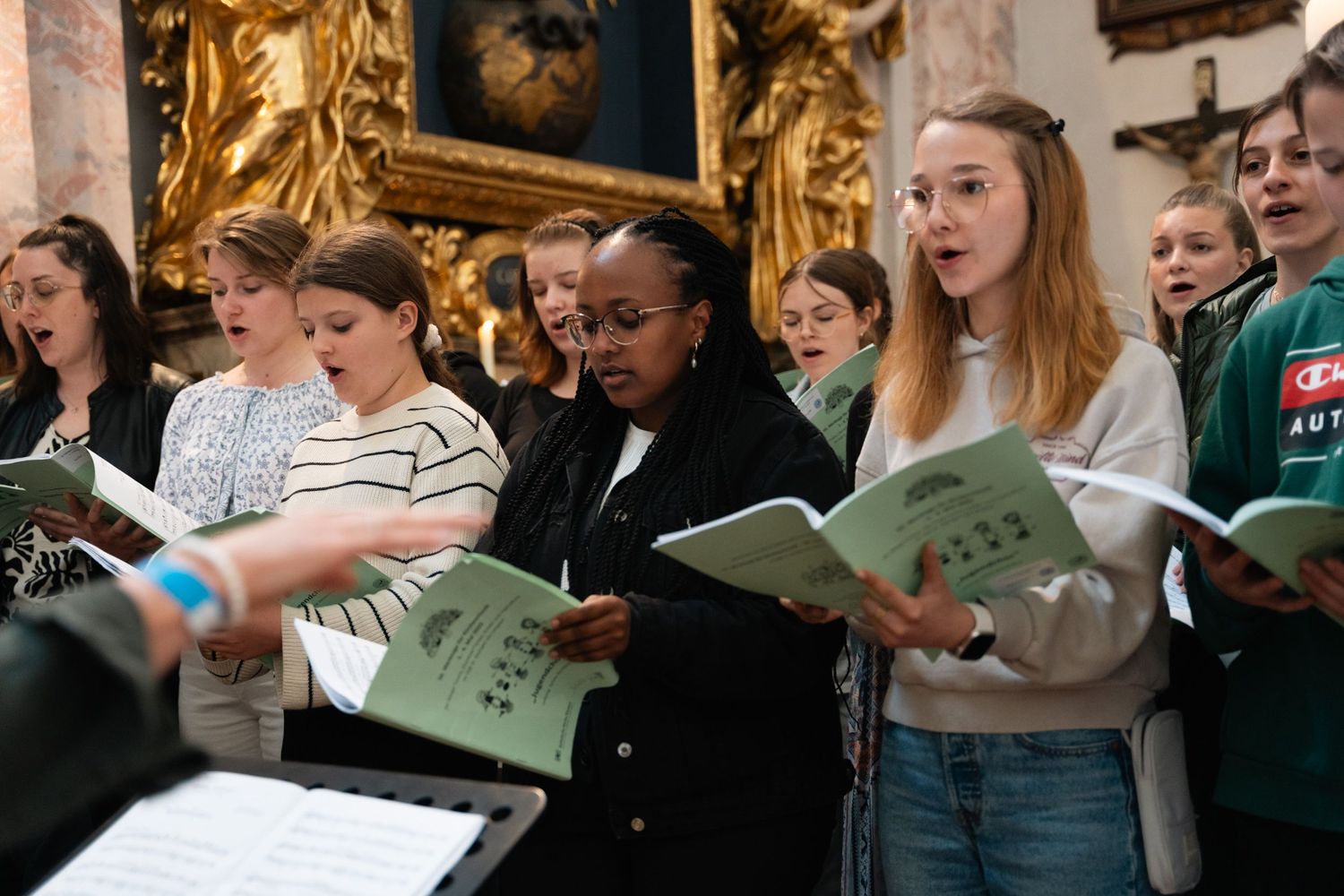 Young choir members singing from green sheet music booklets inside an ornate baroque church.
