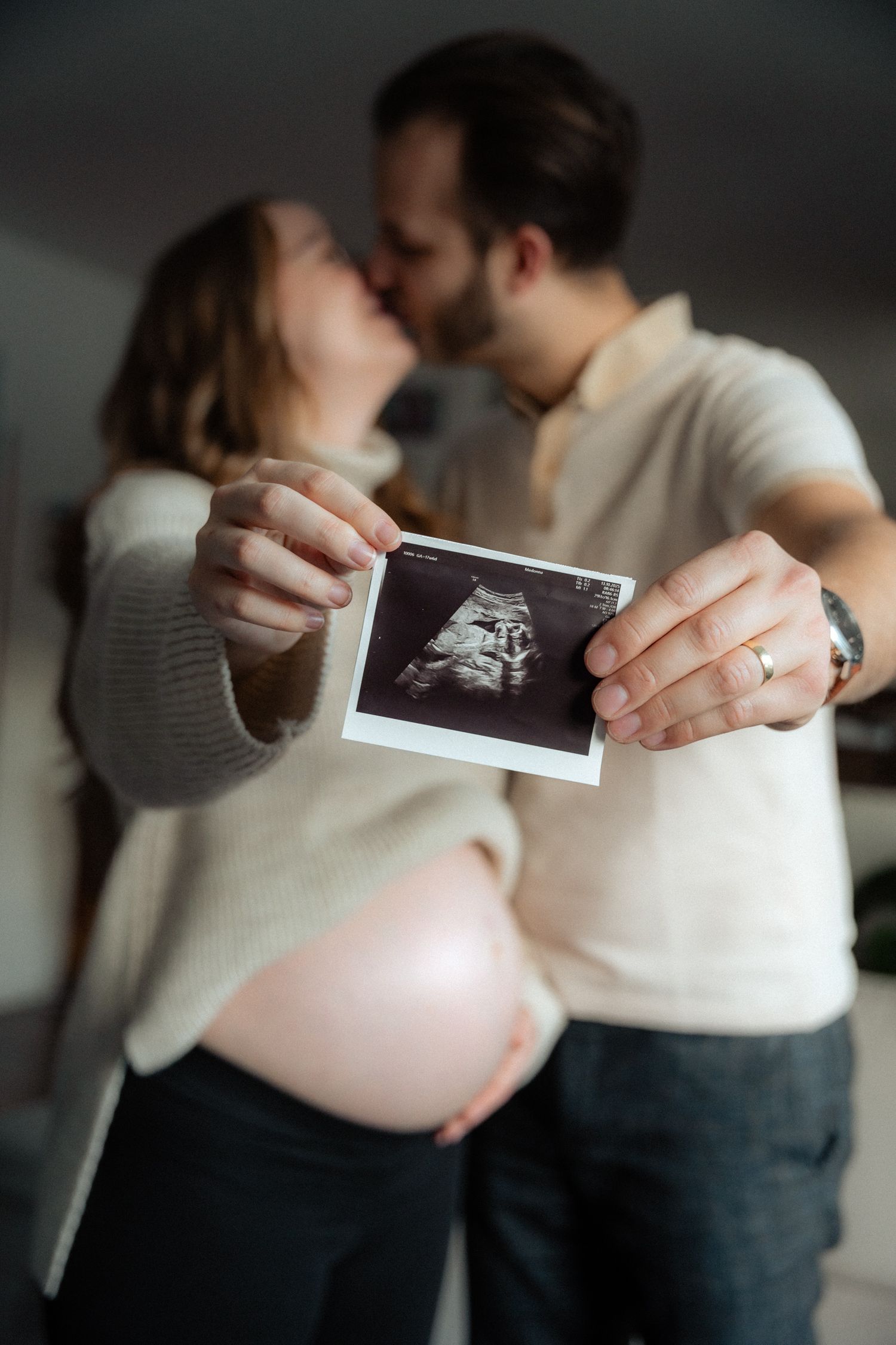 Couple kissing while holding ultrasound photo with pregnant belly visible in foreground.