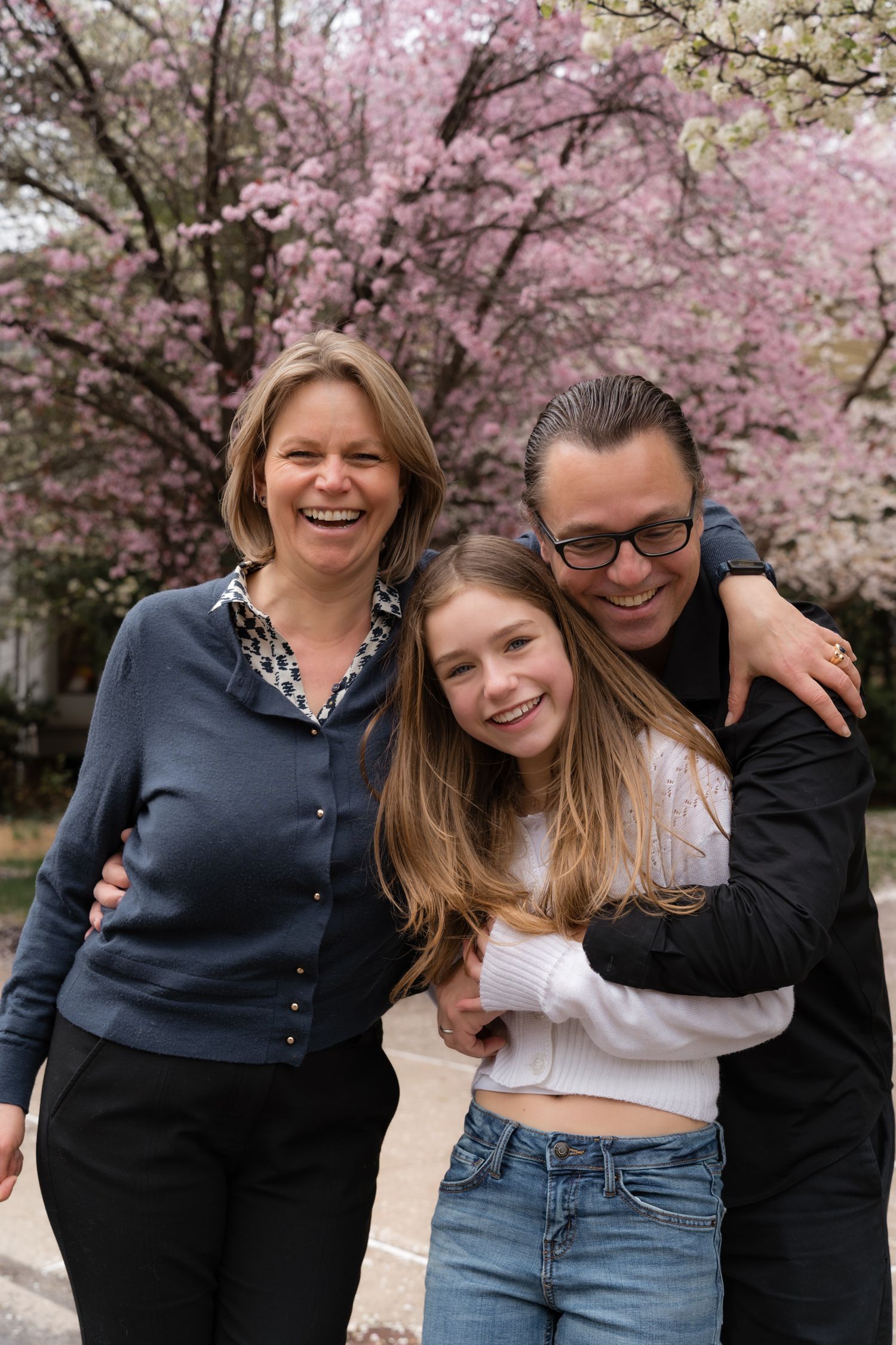 A happy family of three poses together under blooming pink cherry blossom trees in spring.
