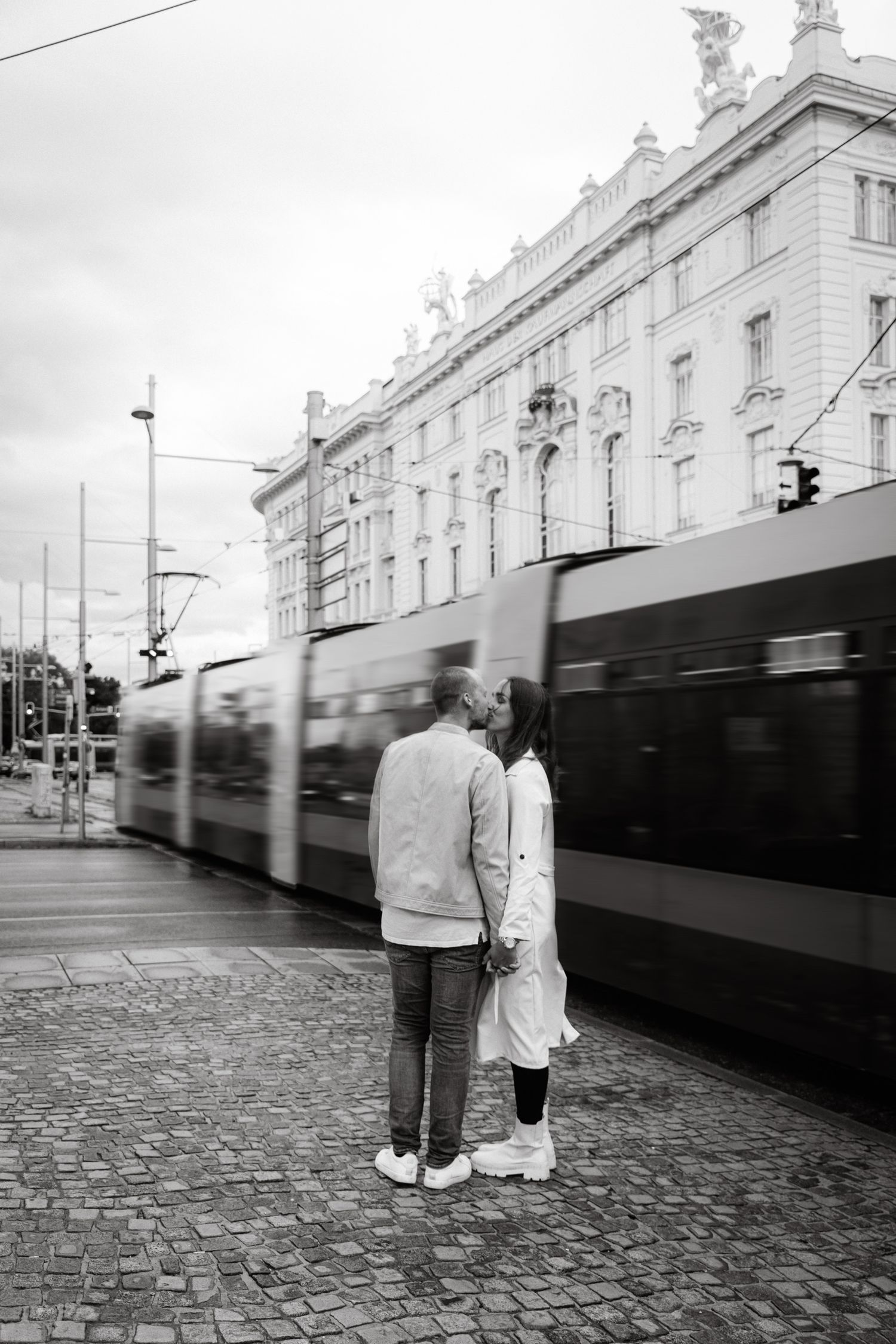 Black and white photo of couple kissing beside a moving tram in a European city square.