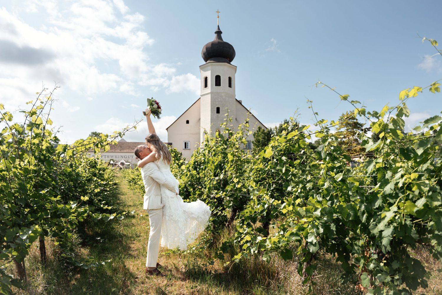 Bride and groom celebrating in a sunny vineyard with a white church steeple in background.