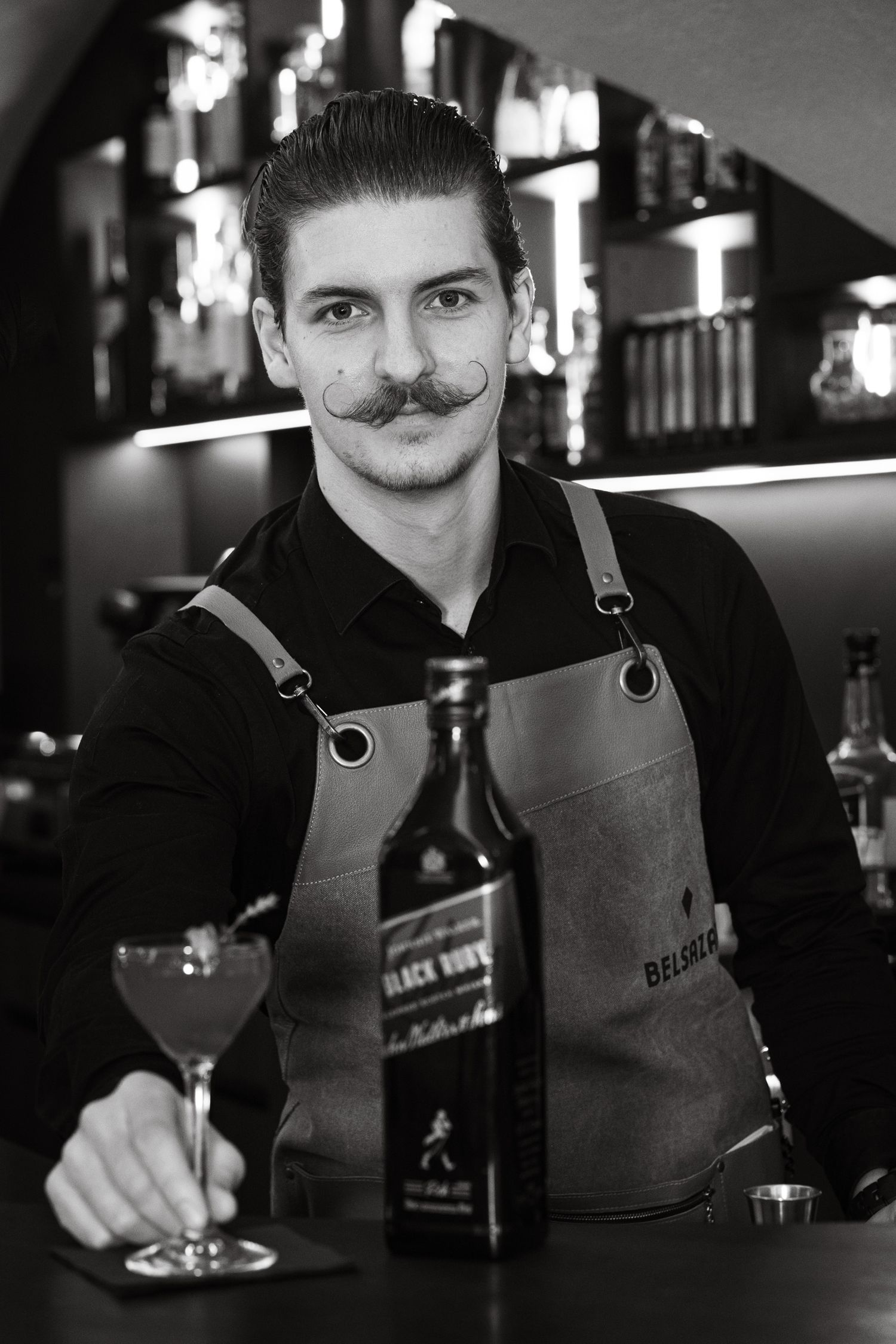 Black and white portrait of a bartender with a mustache posing with Johnnie Walker Black Ruby whisky.