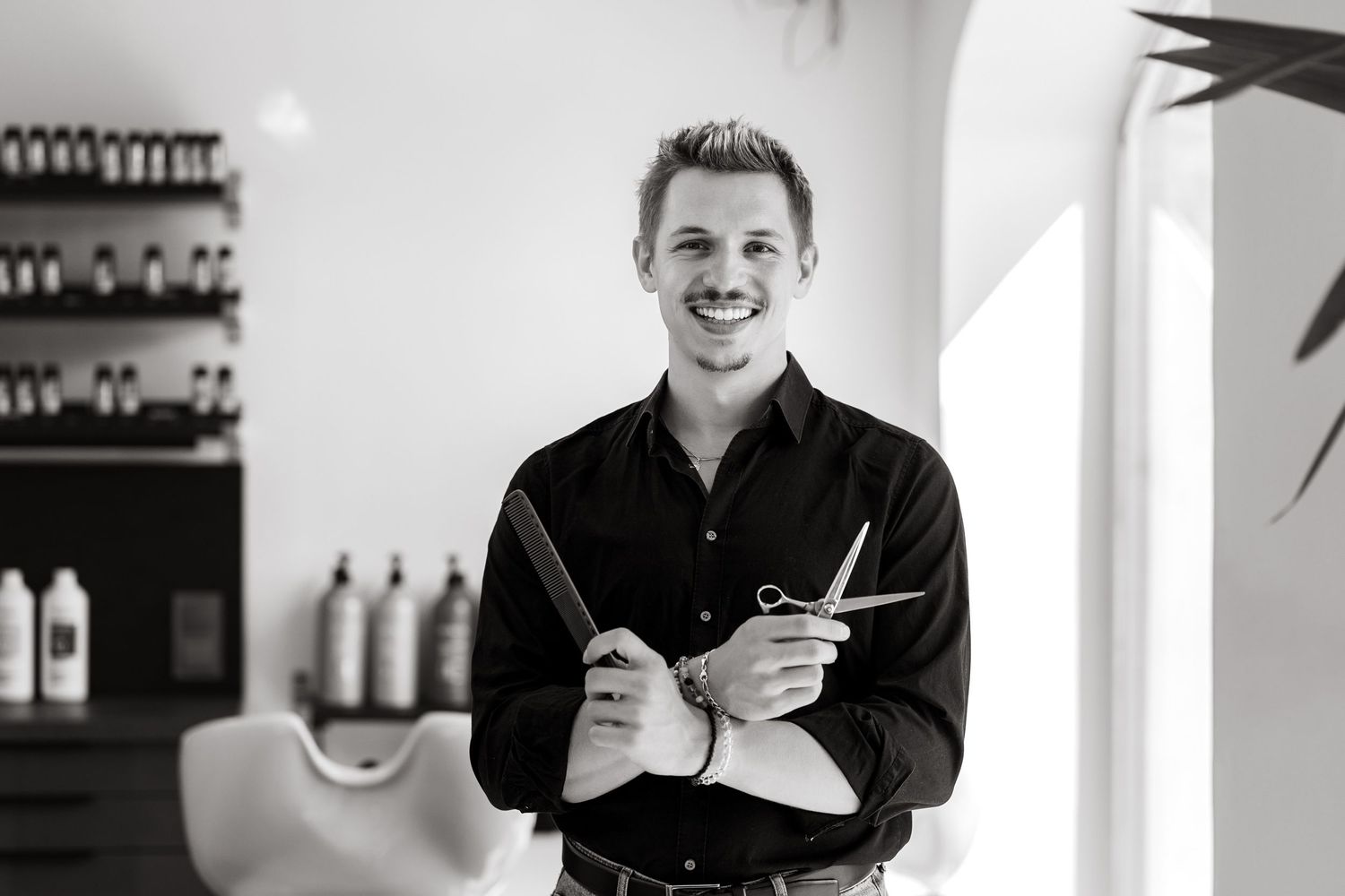 Black and white photo of a smiling male hairstylist holding scissors in a modern hair salon.