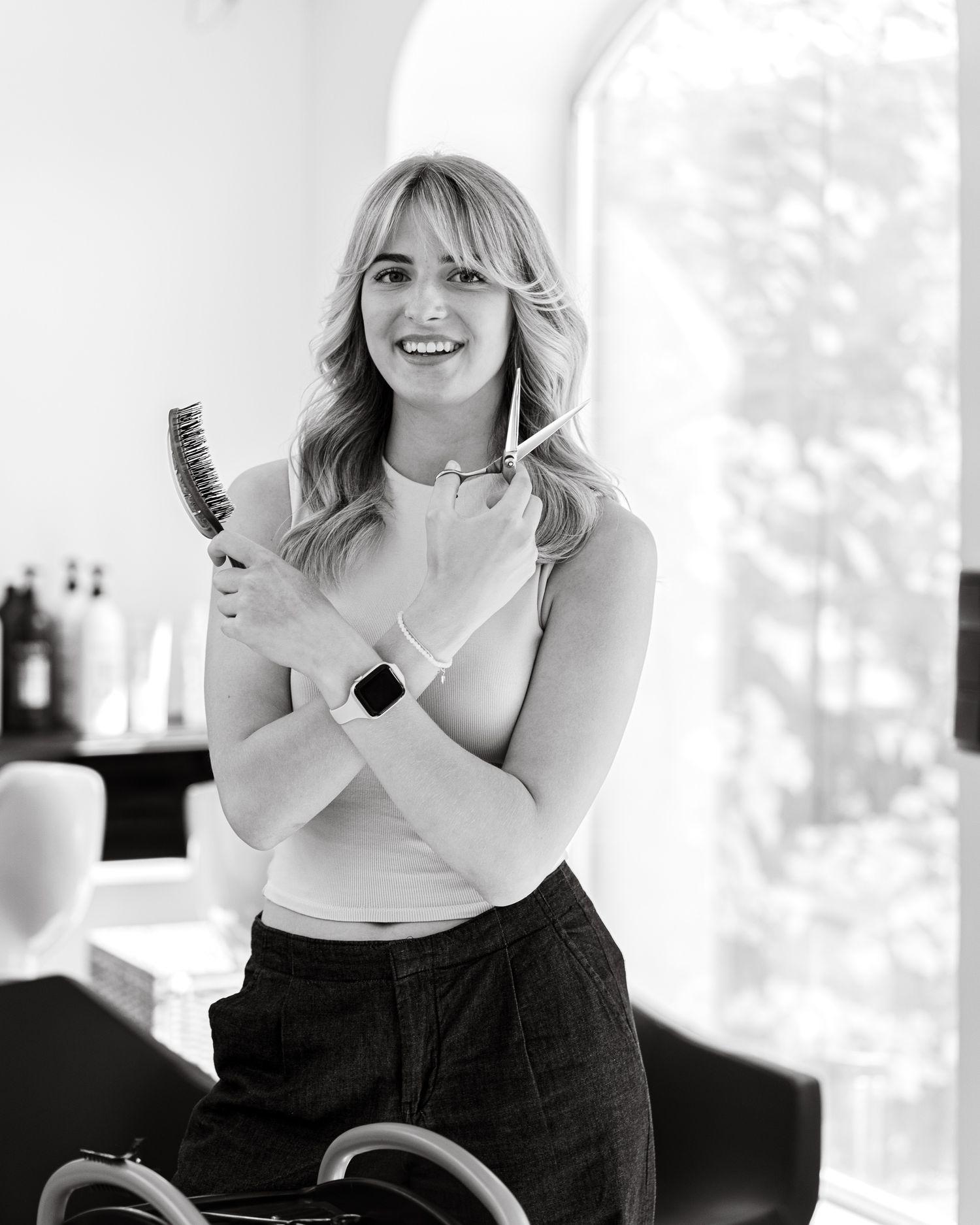 Smiling female hairstylist holding brush and scissors in black and white salon portrait.
