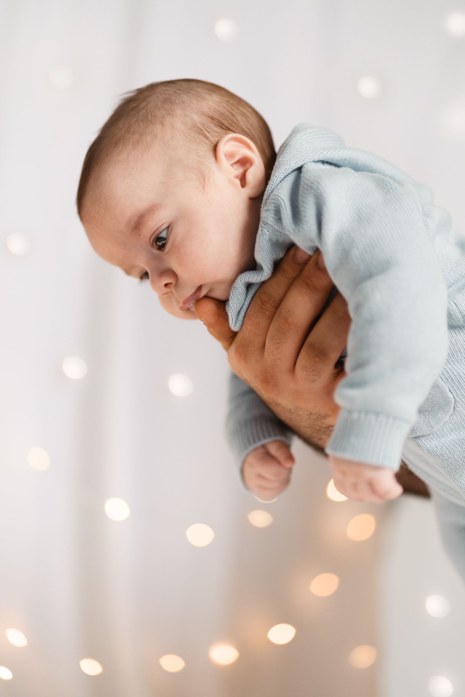 Newborn baby in light blue knit outfit being held against bokeh light background.
