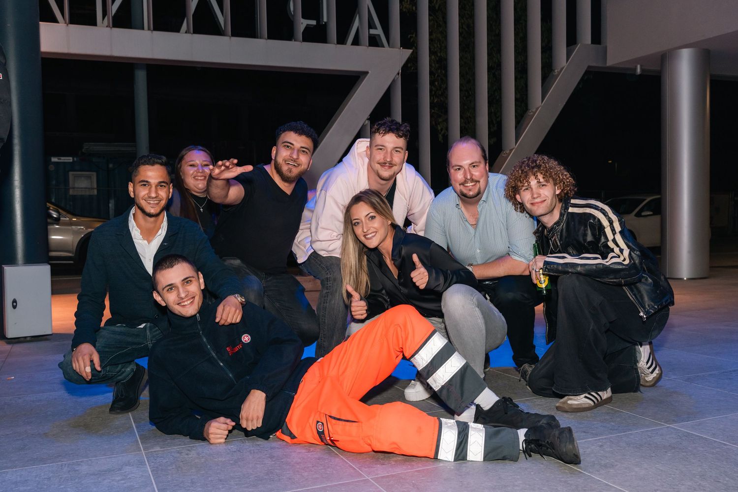 Group of nine young people posing together on a modern outdoor stage at a nighttime event.
