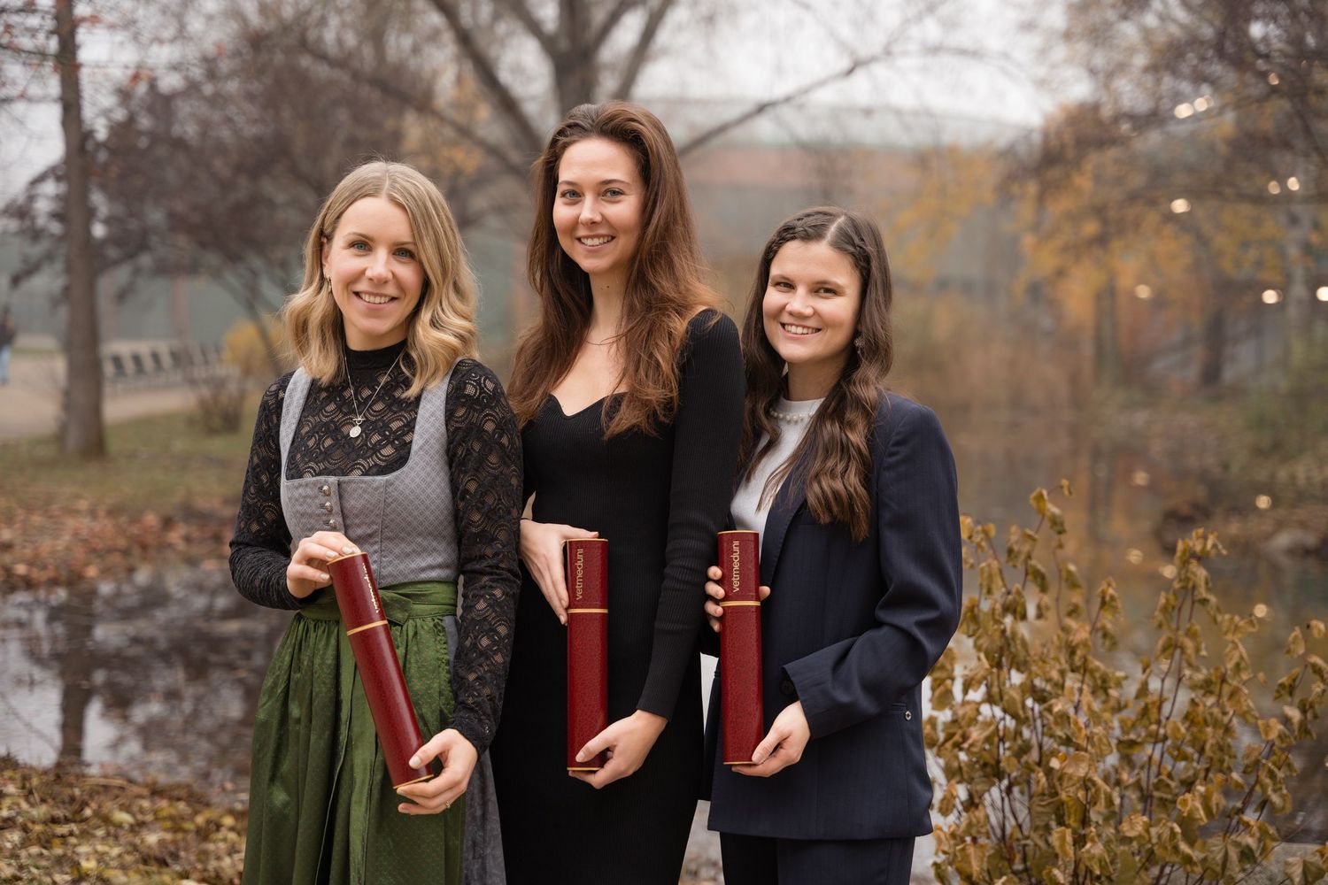 Three smiling women holding diploma tubes posing together outdoors in an autumn landscape.