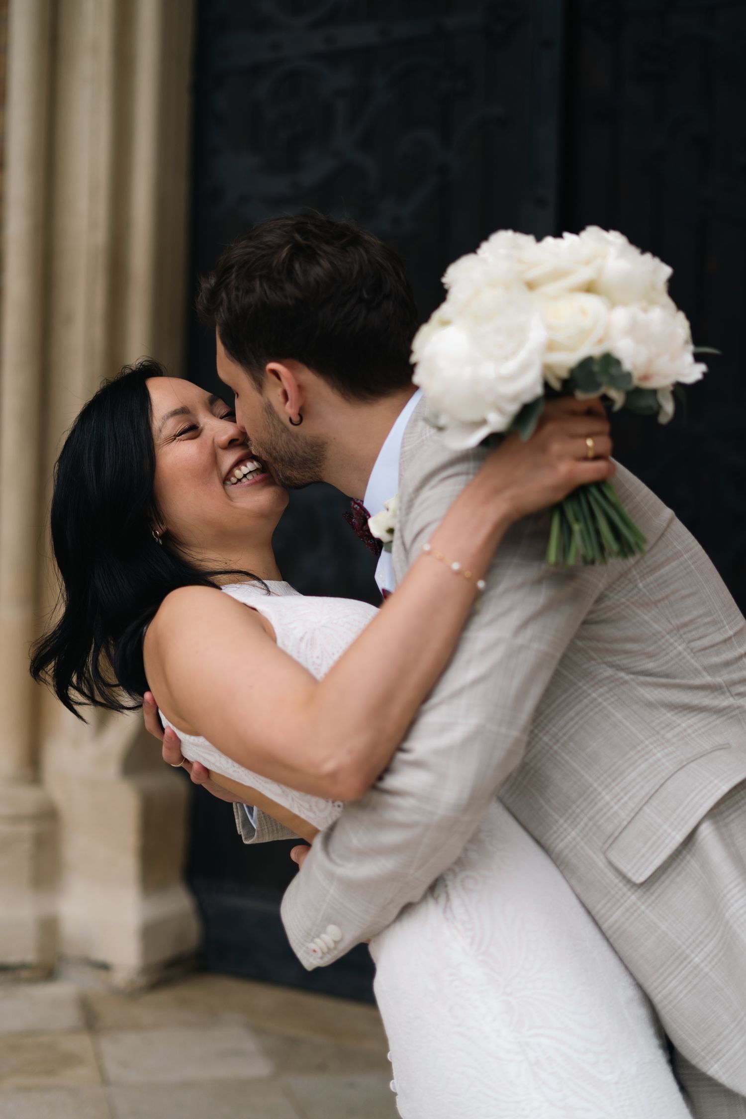 Joyful bride laughing as groom kisses her cheek while holding white peony bouquet outdoors.