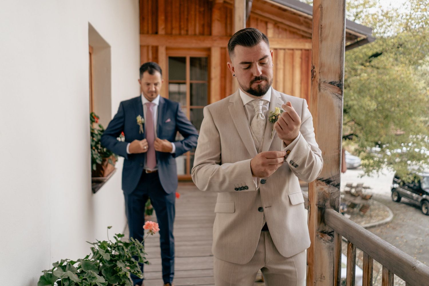 Two groomsmen in suits adjusting cufflinks on a wooden balcony before a wedding ceremony.