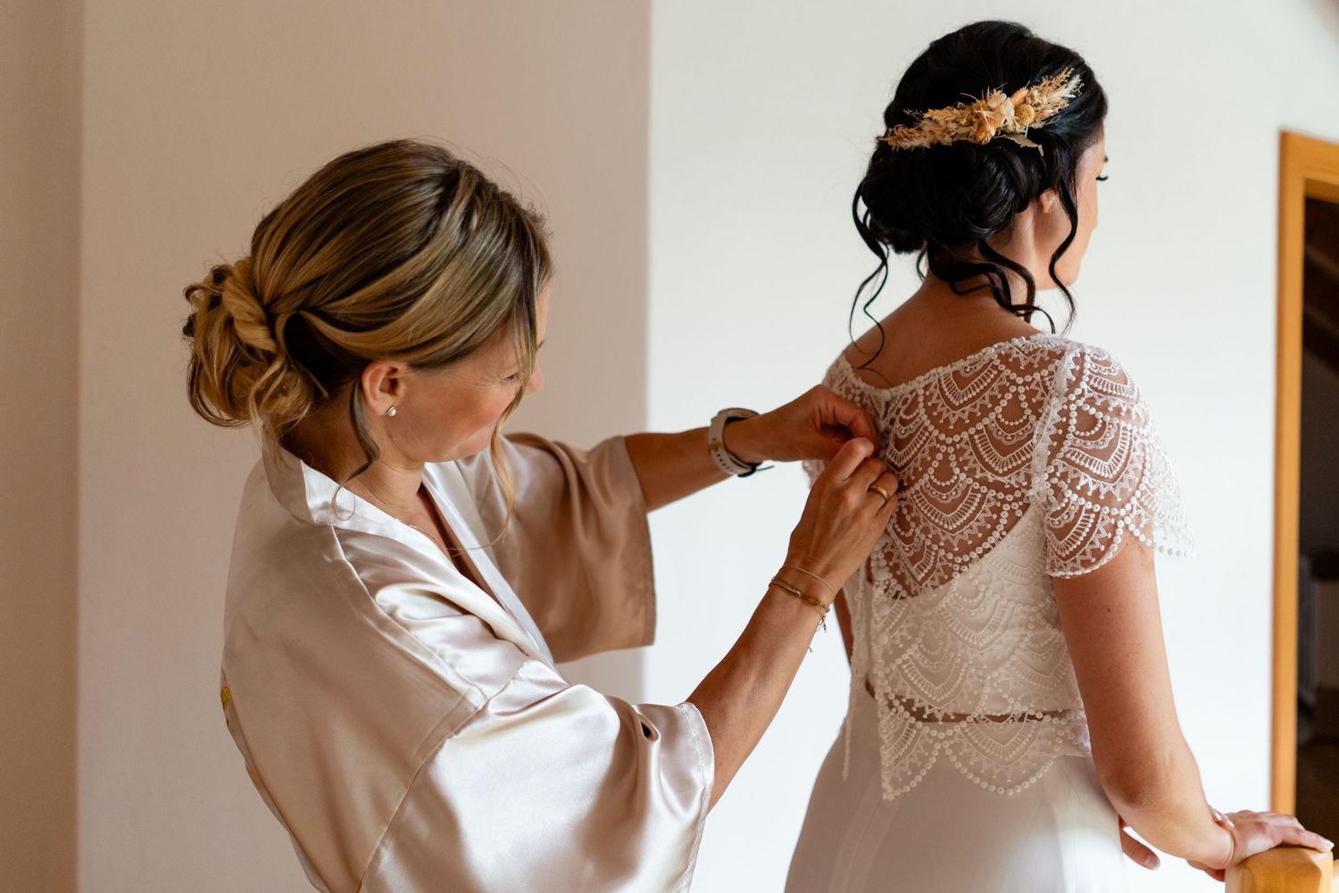 Woman in satin robe fastening intricate lace wedding dress on bride with gold hair piece.