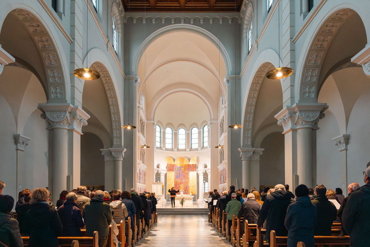 Interior of a grand church with arched columns and congregation seated in wooden pews during a service.