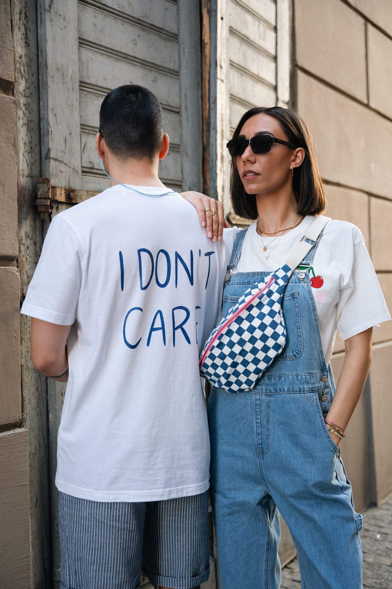 Street style couple, woman in denim overalls with checkered bag, man in white tee.