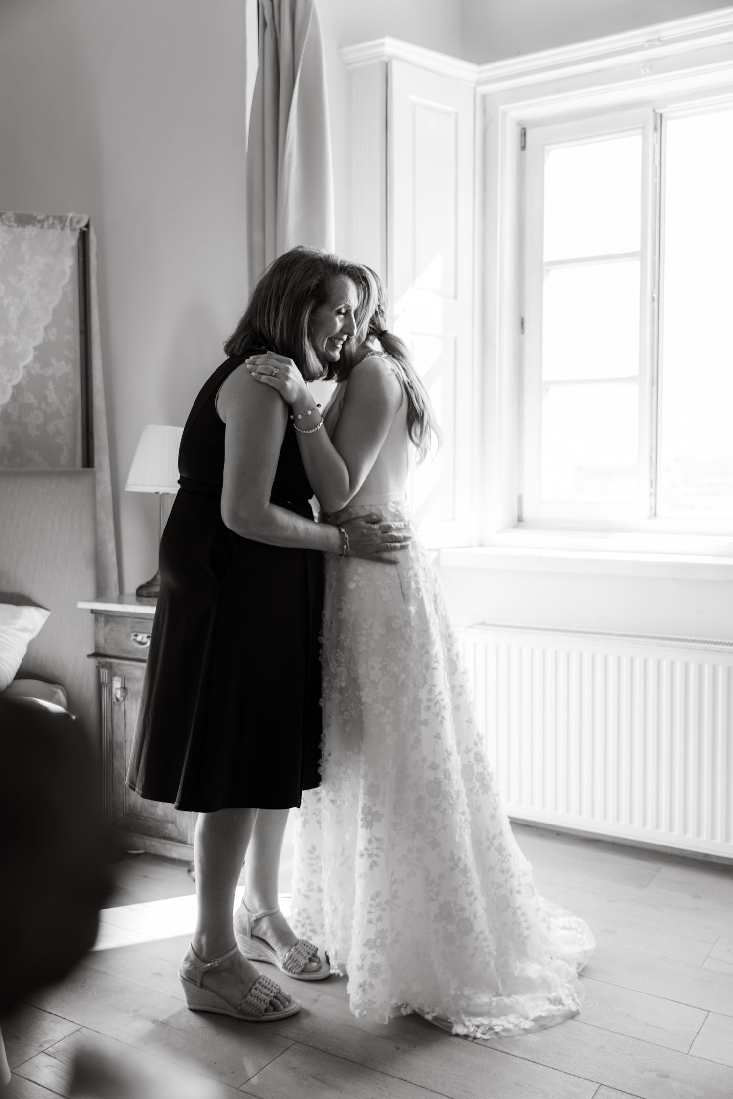 Black and white photo of bride and mother sharing an emotional embrace by a bright window.