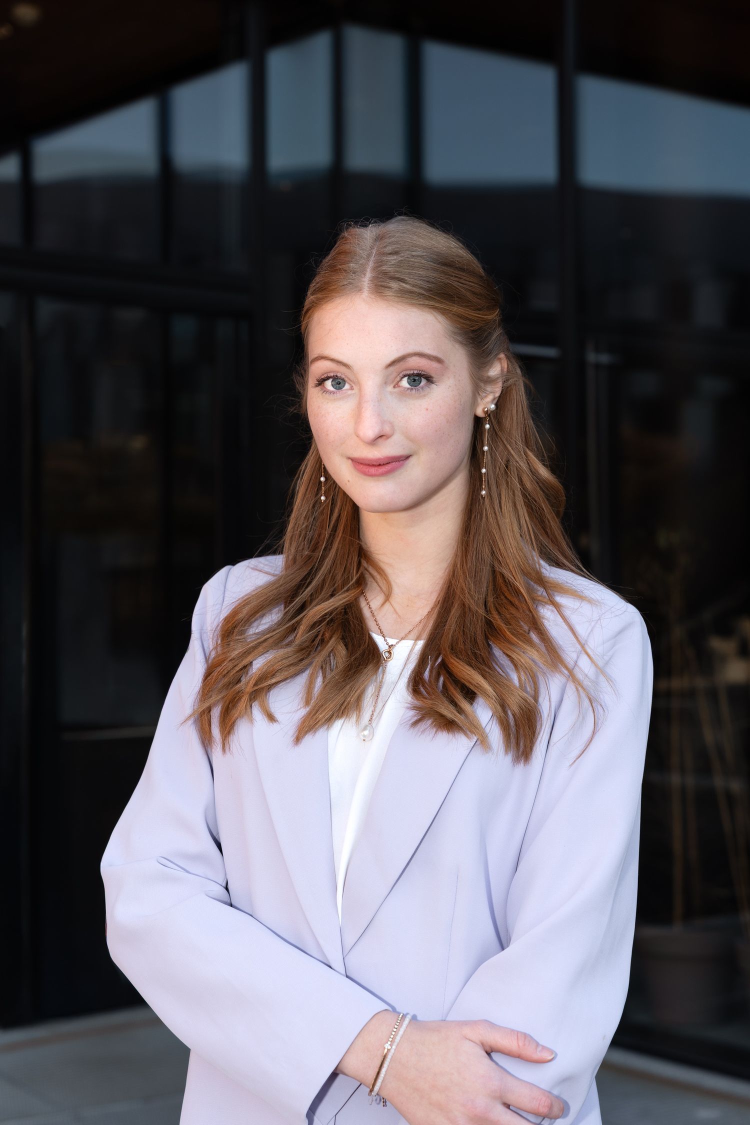 Young professional woman in lavender blazer with long auburn hair posing outdoors in front of modern building.