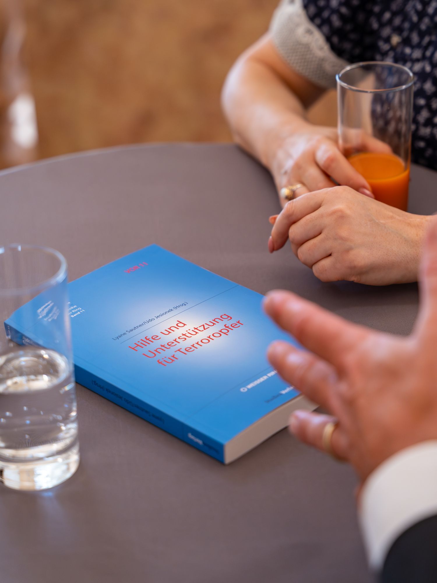 Blue book titled Hilfe und Unterstützung der Terroropfer on a round table with glasses of water and juice.
