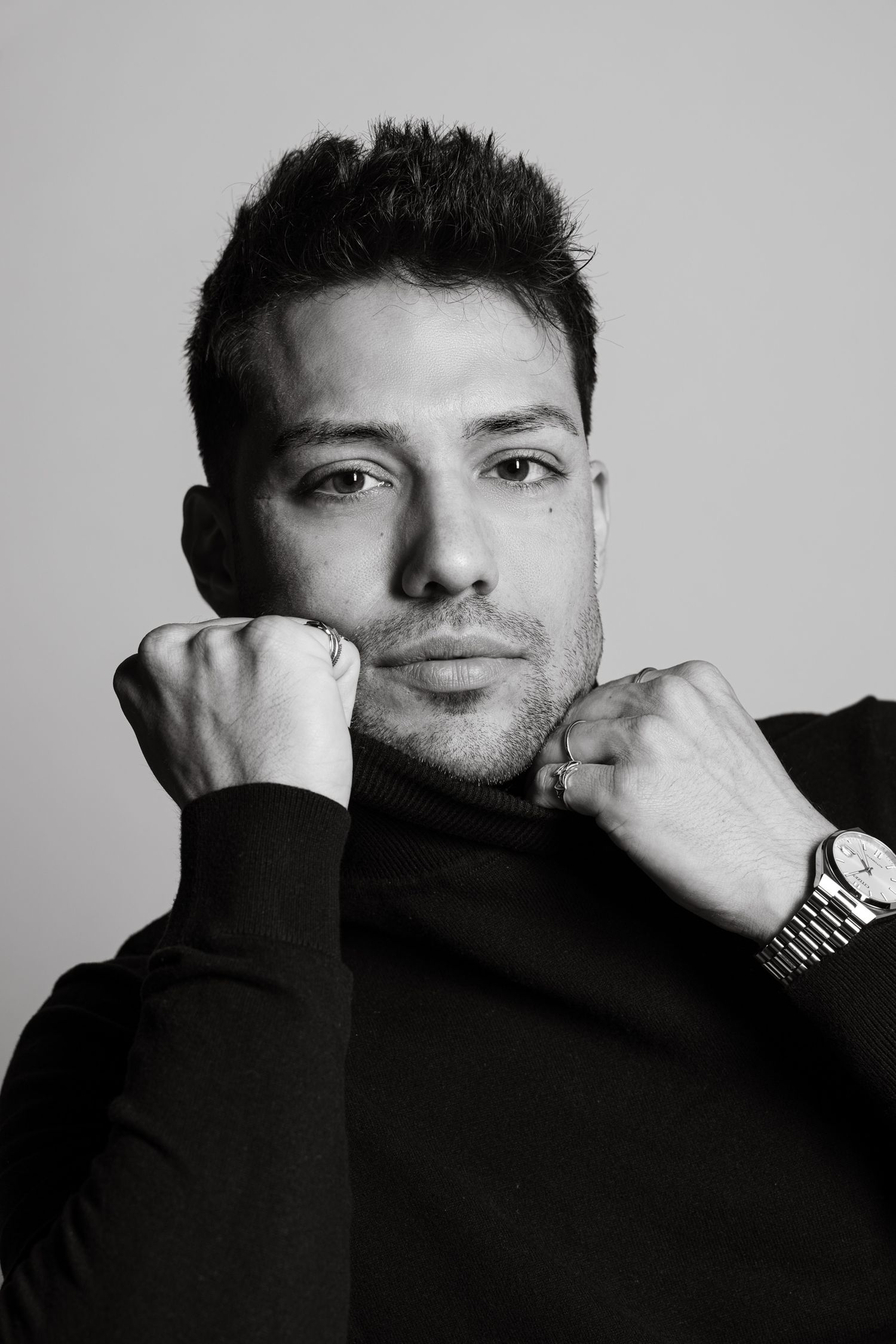 Black and white portrait of a man in a turtleneck wearing a watch, posing with hands raised to chin.