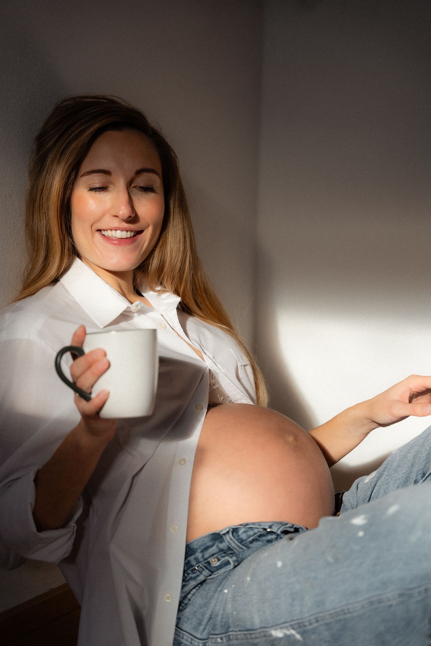Smiling pregnant woman in white shirt and jeans holding a white mug, enjoying warm sunlight indoors.