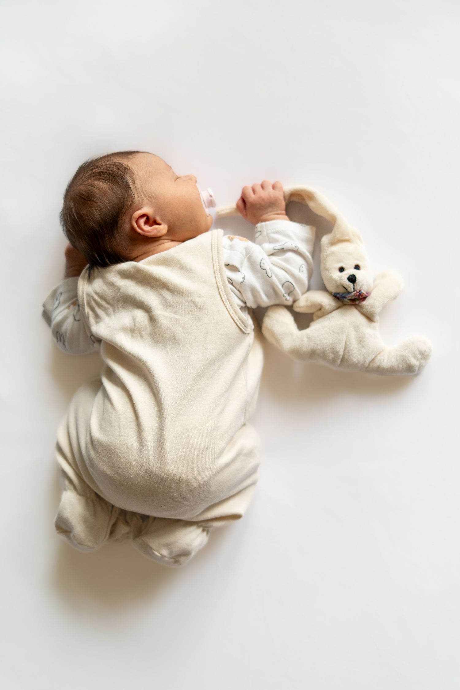 Newborn baby sleeping peacefully in white onesie with a soft teddy bear toy.