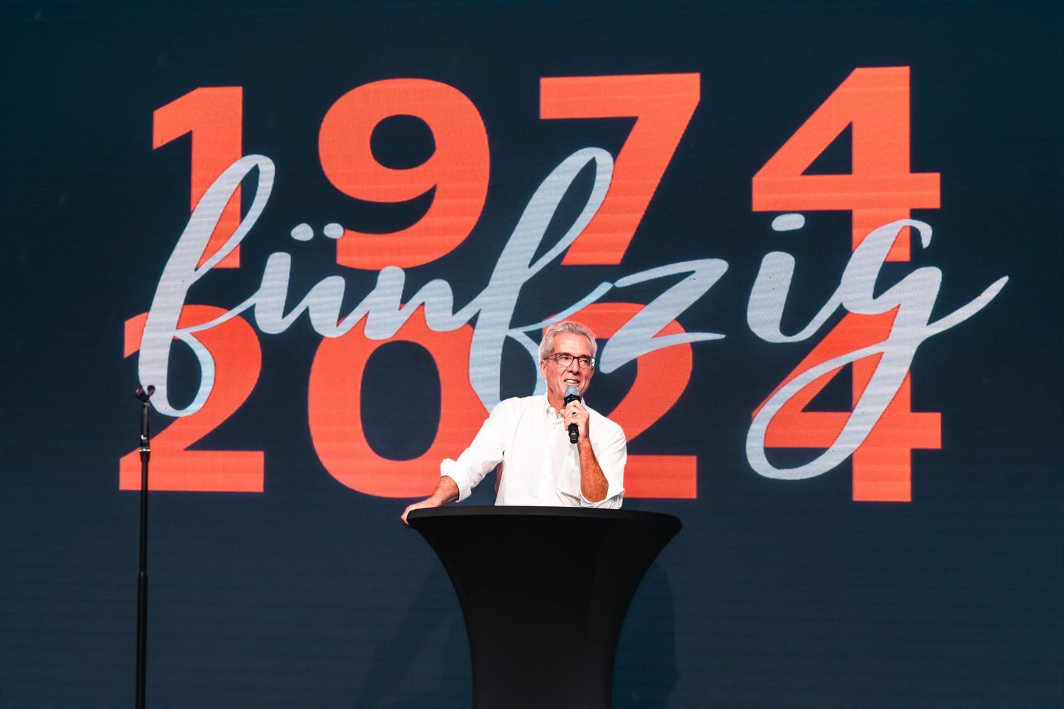 Speaker at podium presenting in front of large screen displaying 1974-2024 fünfzig 50th anniversary backdrop.
