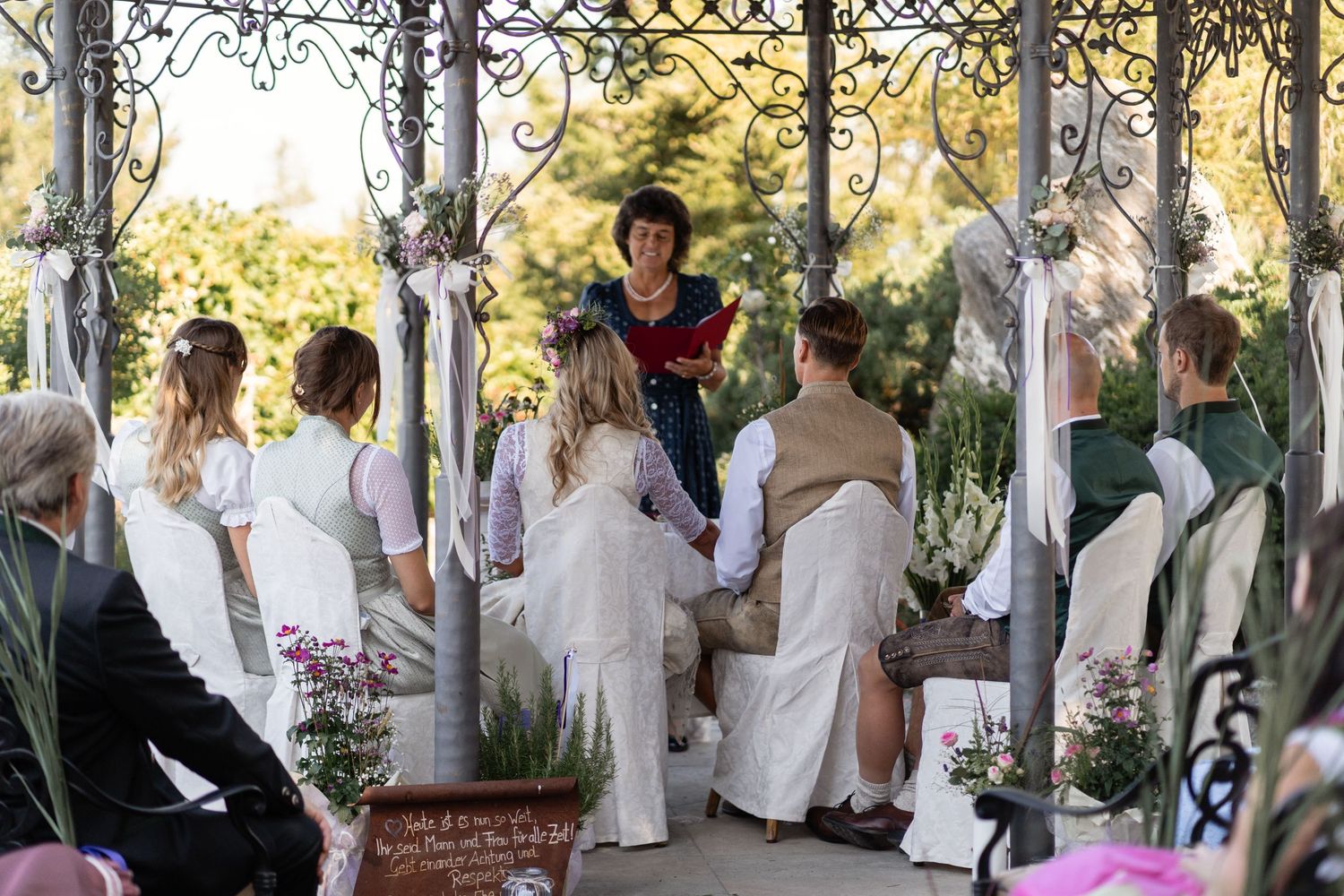 Outdoor garden wedding ceremony with couple holding hands before officiant under ornate iron arbor.