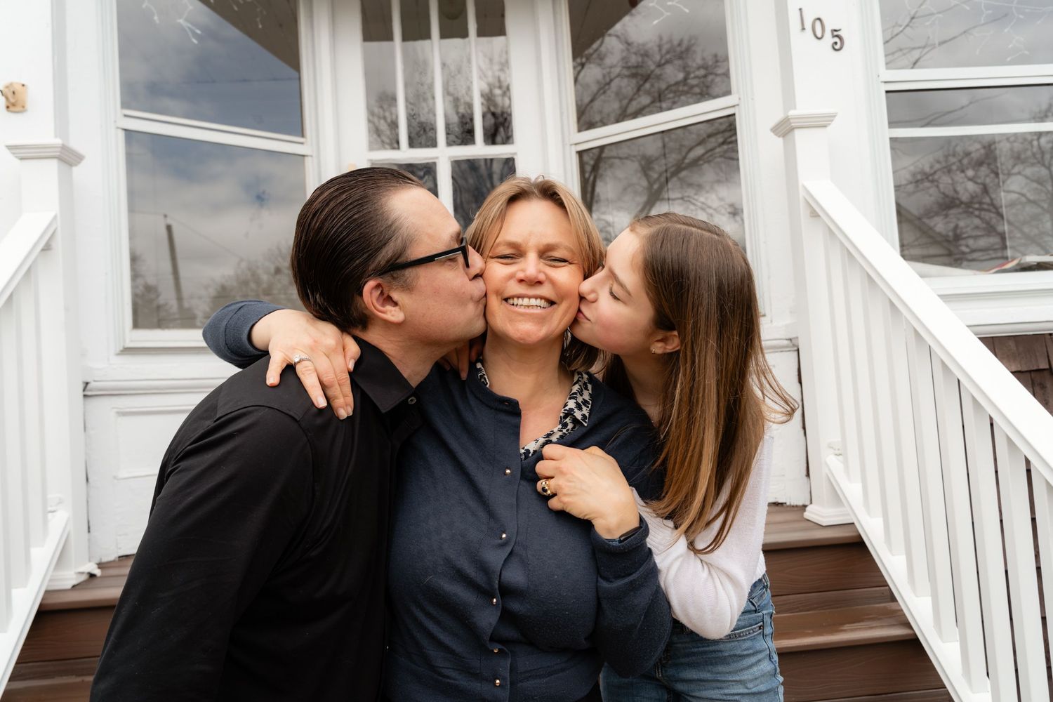 A happy family of three sharing kisses on the front porch of a white house.