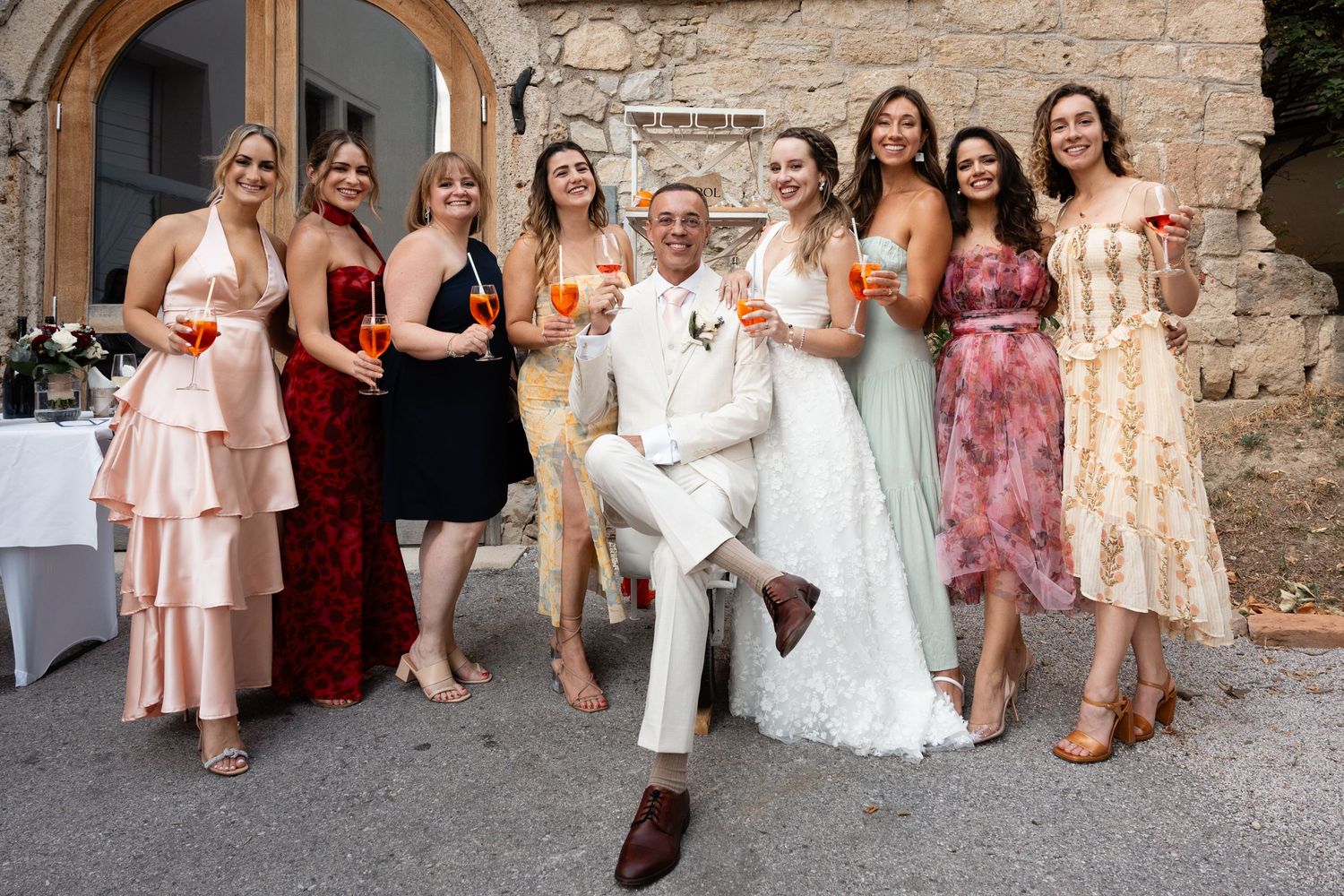 Groom in white suit poses with bride and colorful bridesmaids holding drinks.