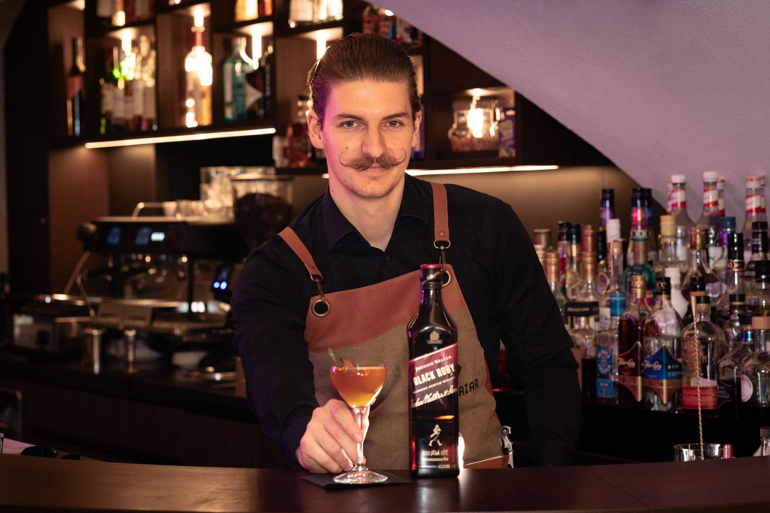 Smiling bartender in apron holding a cocktail glass beside a Jack Daniel's bottle at a stylish bar.