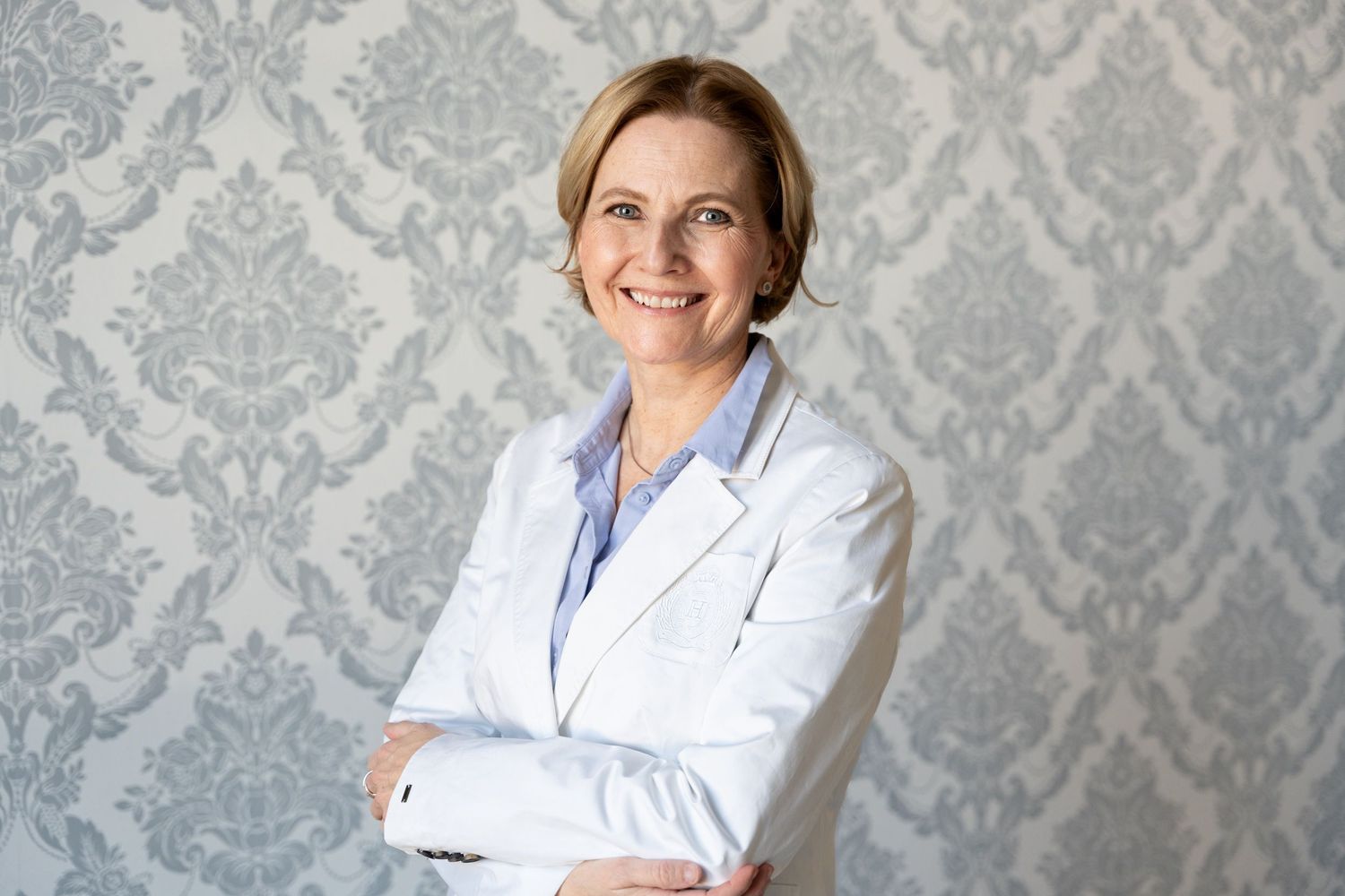 Smiling female doctor in white lab coat standing with arms crossed against decorative wallpaper.