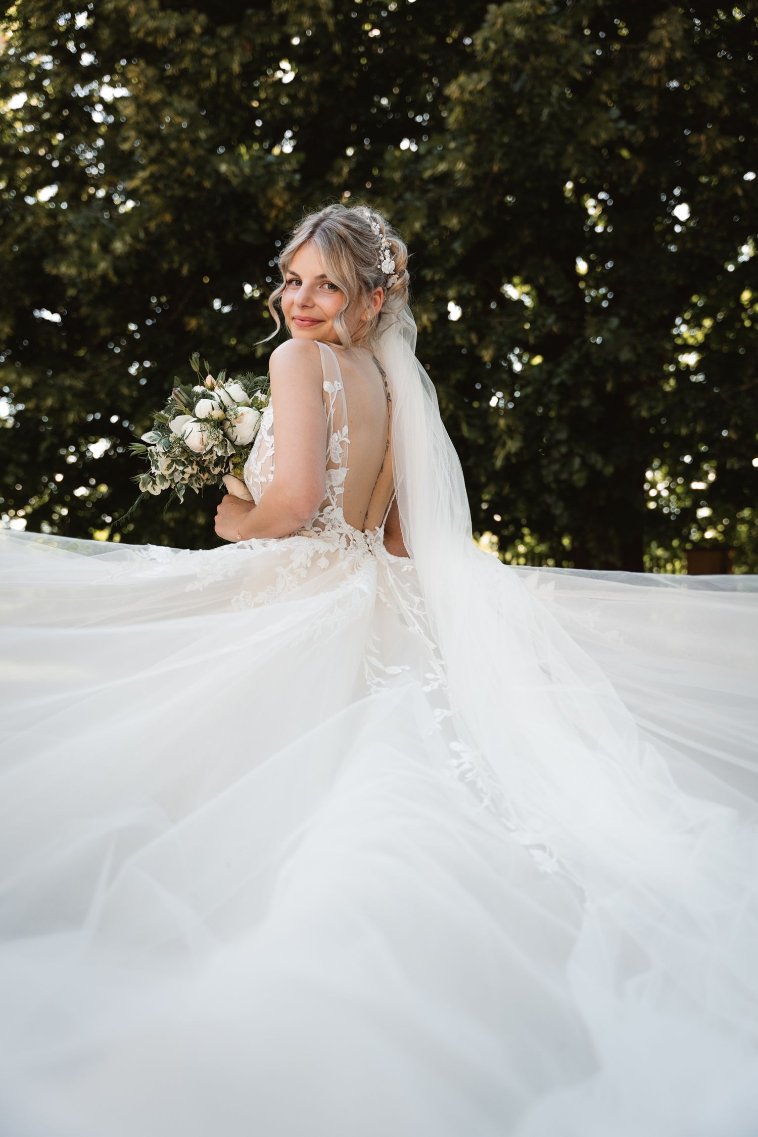 Bride in backless lace wedding gown with flowing veil holds bouquet outdoors.