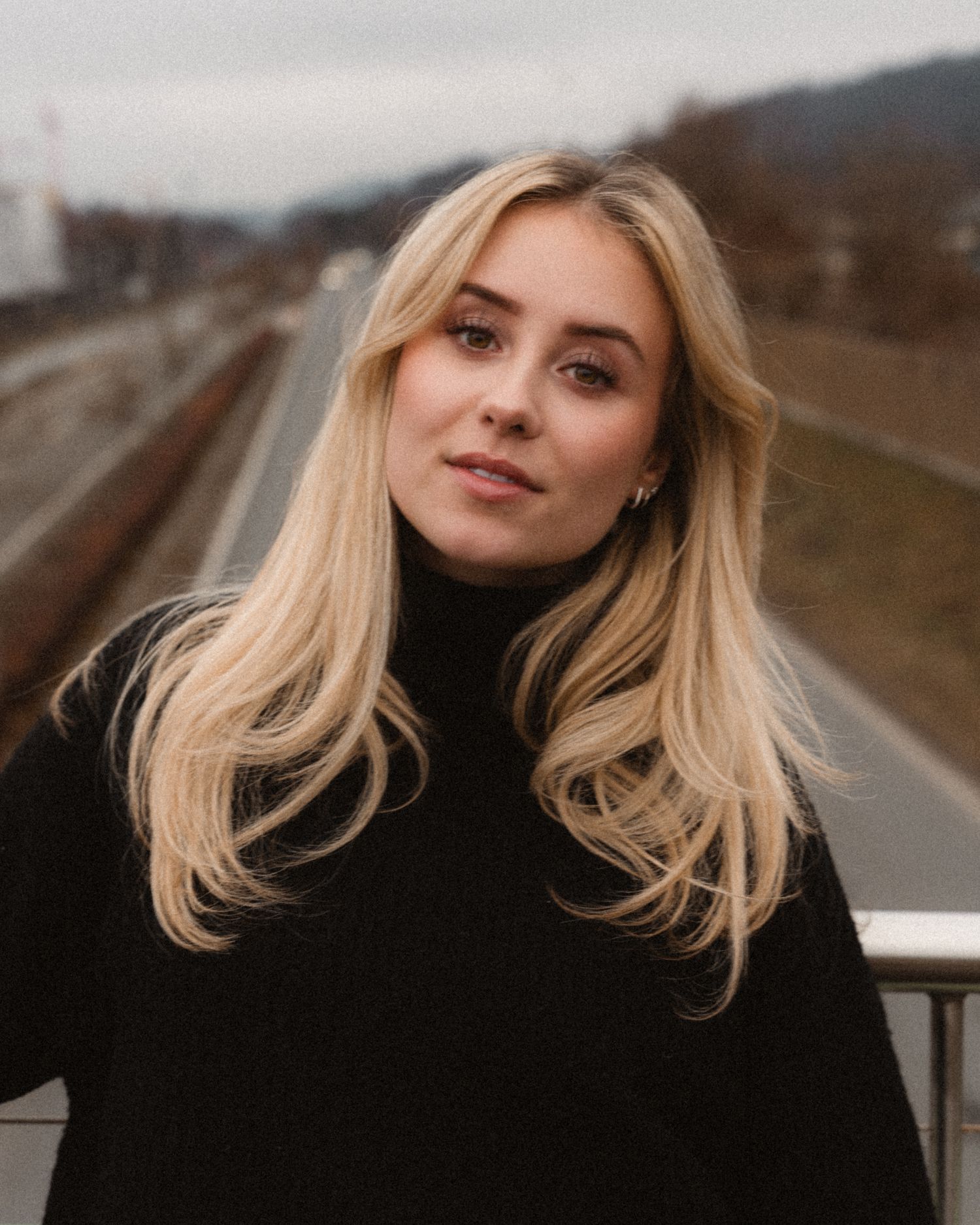 Blonde woman in black turtleneck sweater posing on bridge with railway tracks behind her.