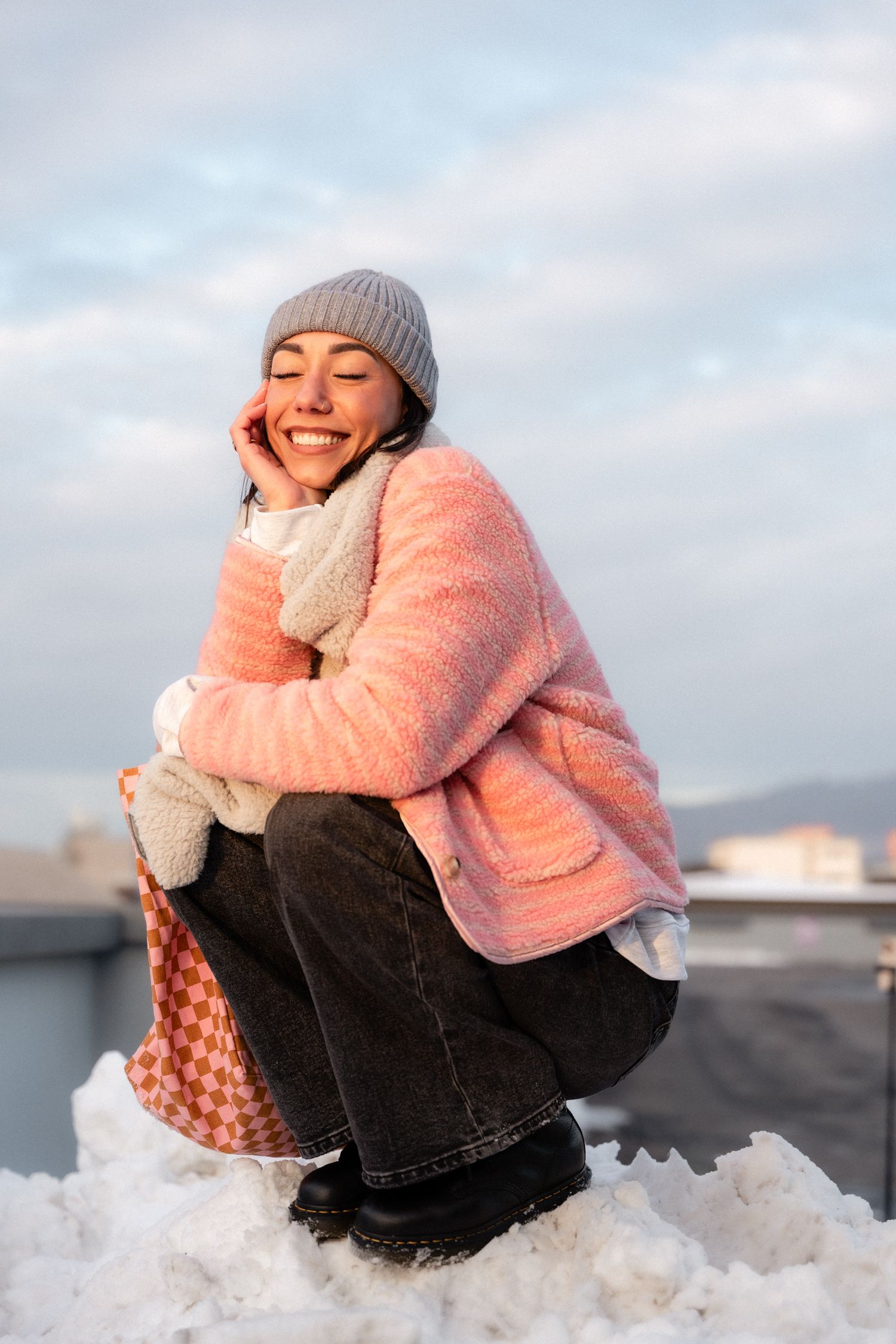 Happy woman in a pink fluffy jacket and grey beanie crouching on snow under a cloudy sky.