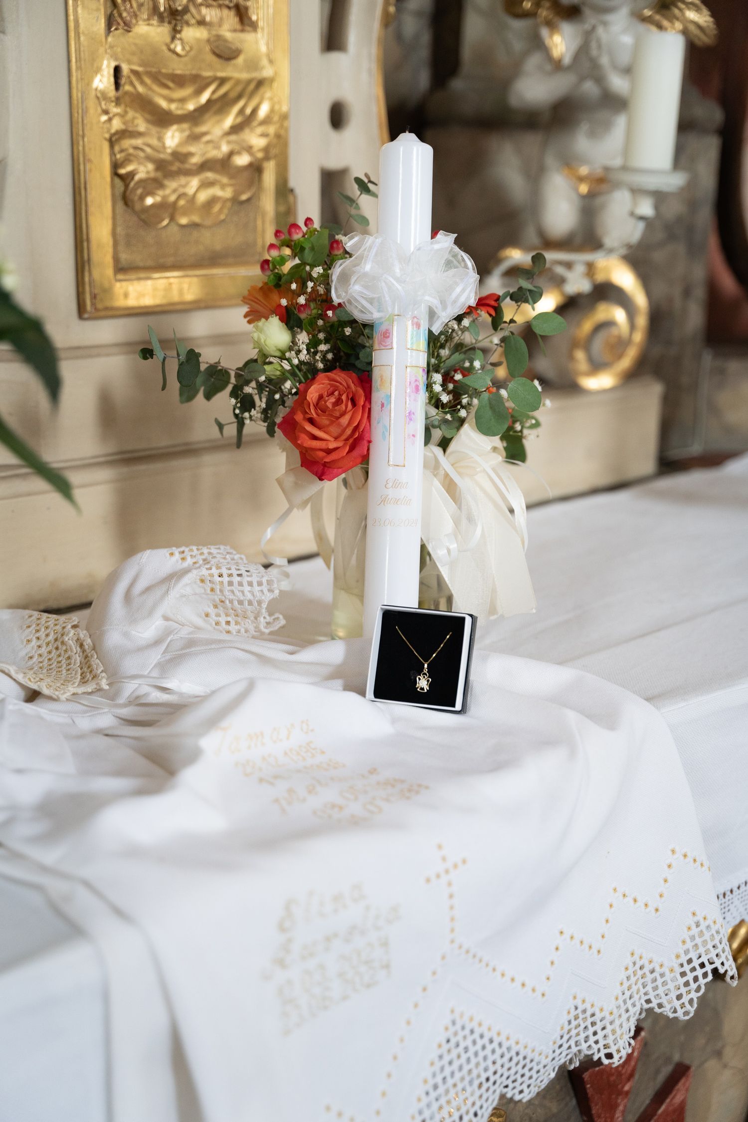 A white baptism candle decorated with red roses and a cross necklace on an embroidered cloth.