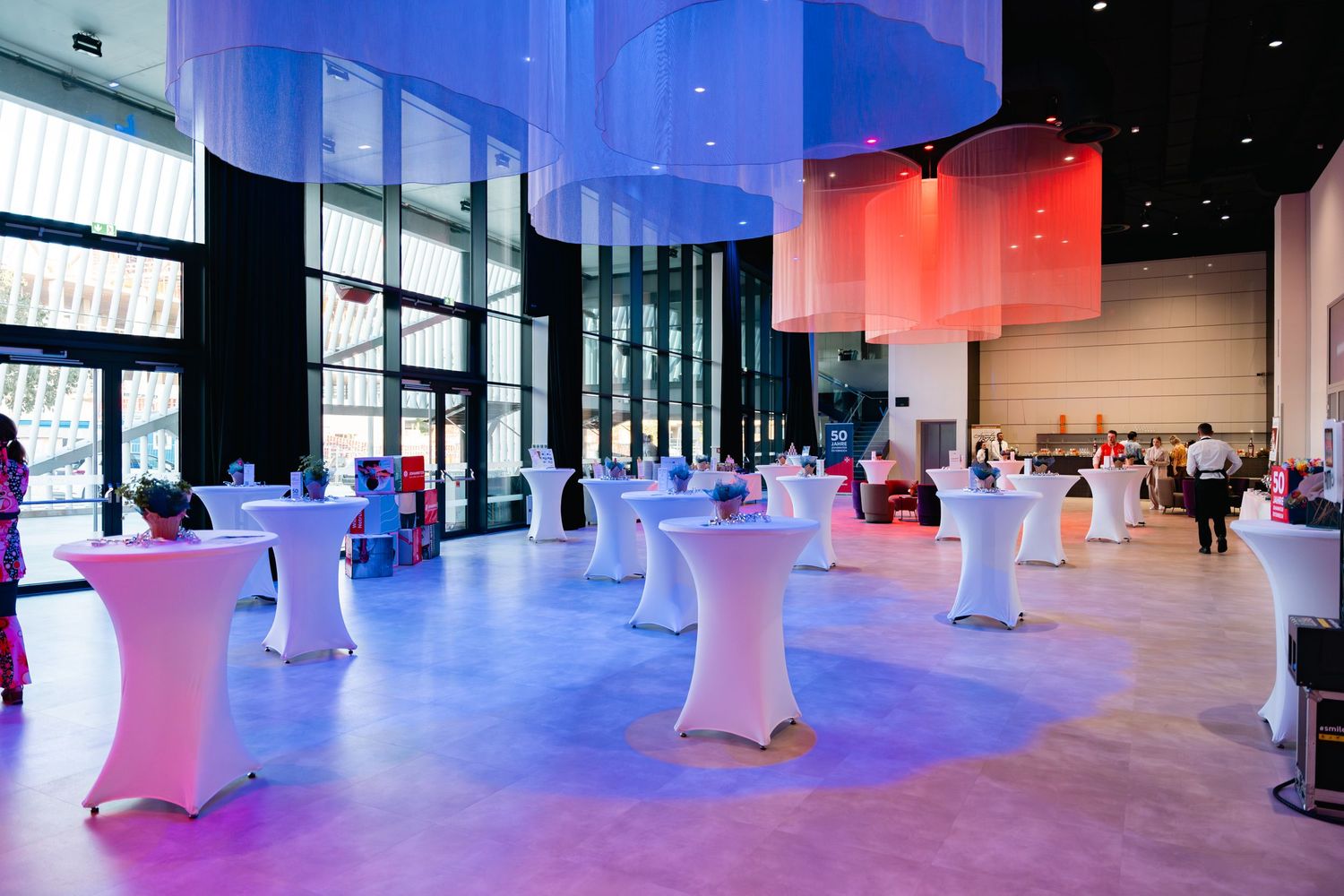 Elegant event venue with white cocktail tables, colorful hanging decorations, and vibrant purple and blue lighting.
